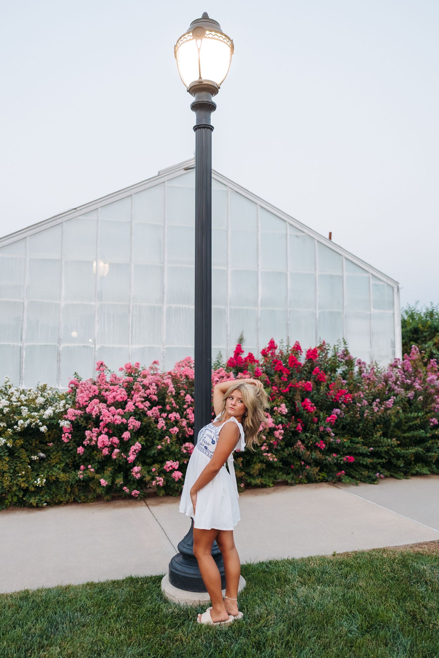 Blonde woman in white dress posing by a lit lamp post with pink flowering bushes and greenhouse backdrop.