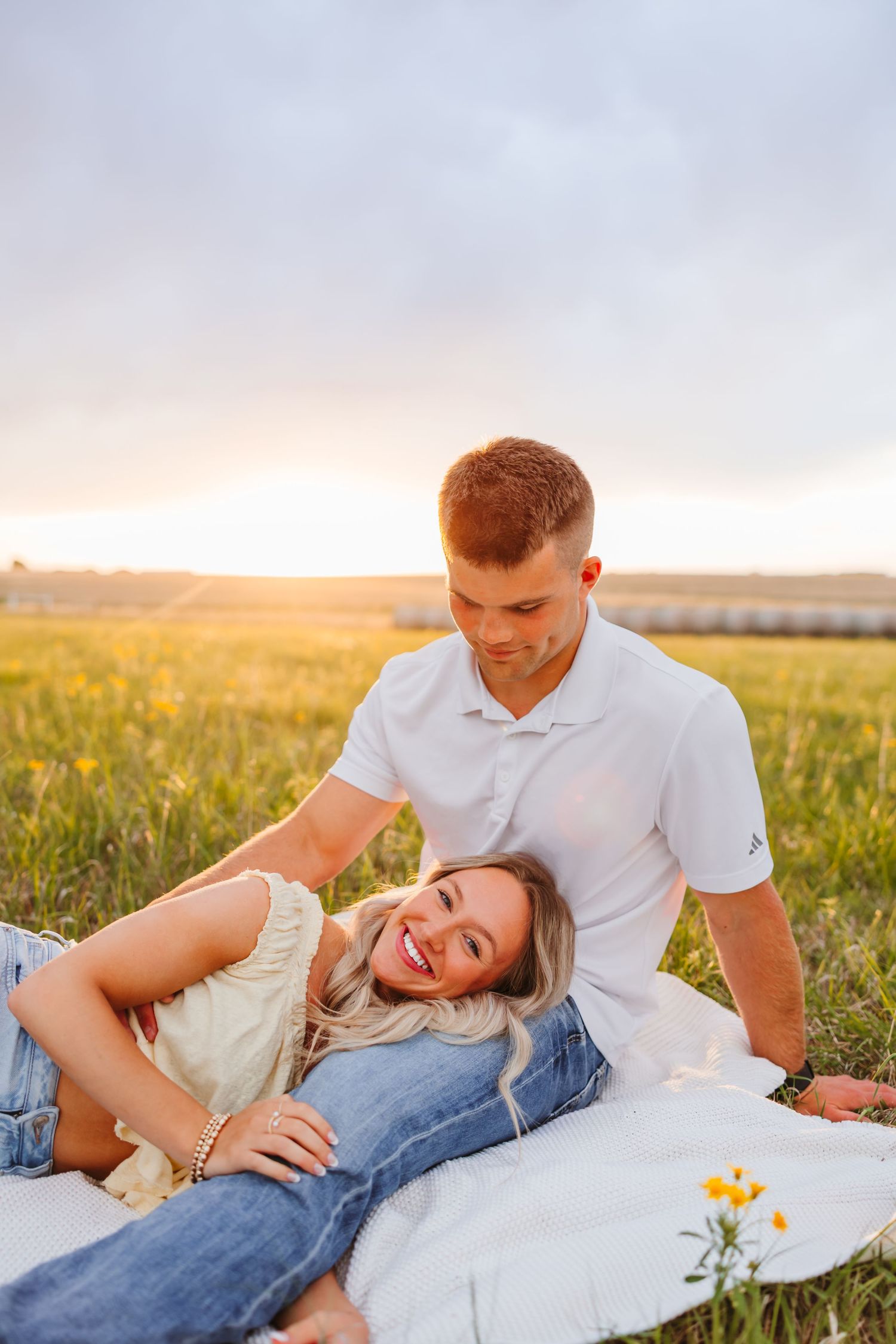 Couple relaxing in a golden field at sunset, woman lying on blanket smiling up at man in white polo shirt.