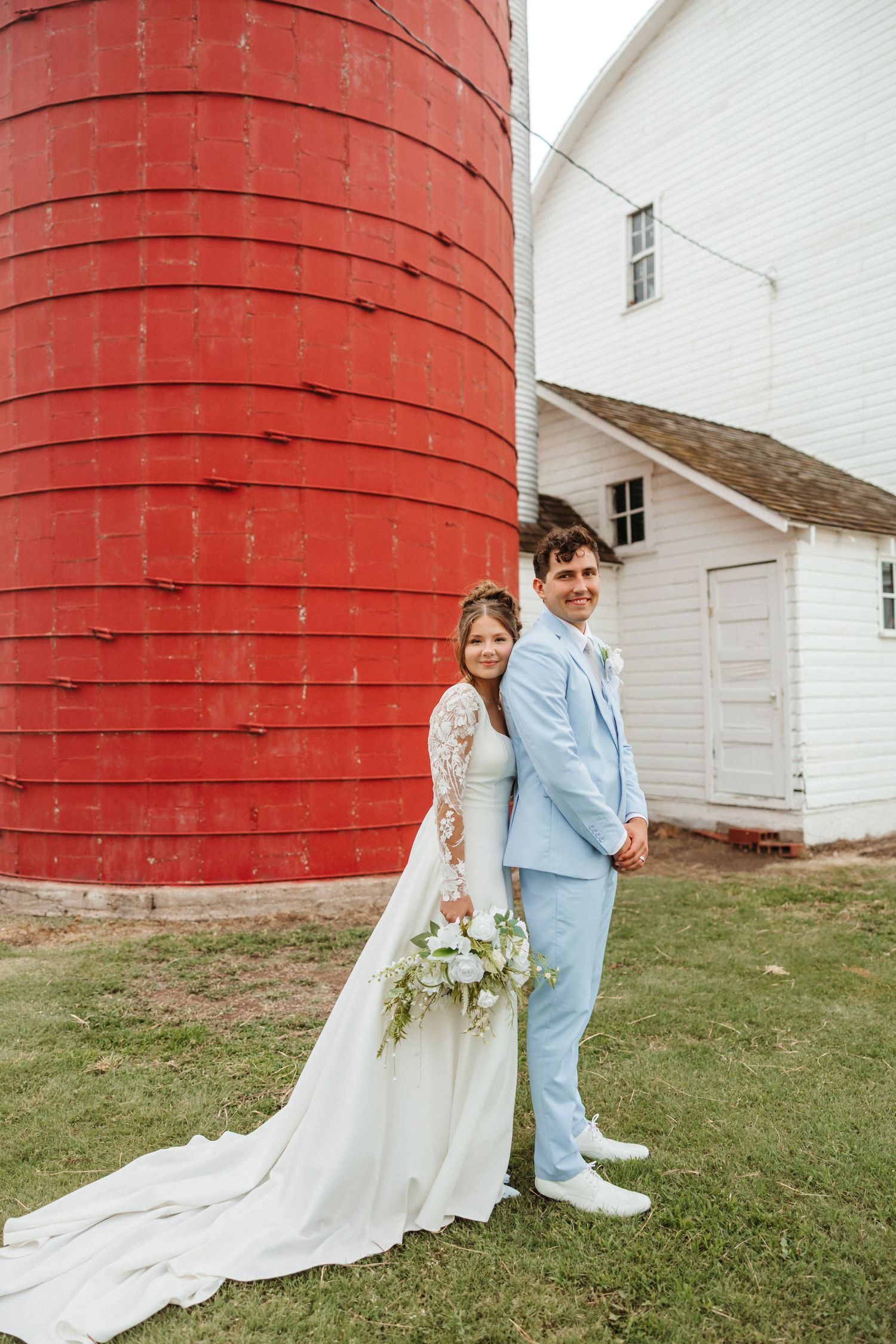 Bride and groom pose together in front of a red silo and white barn on their wedding day.