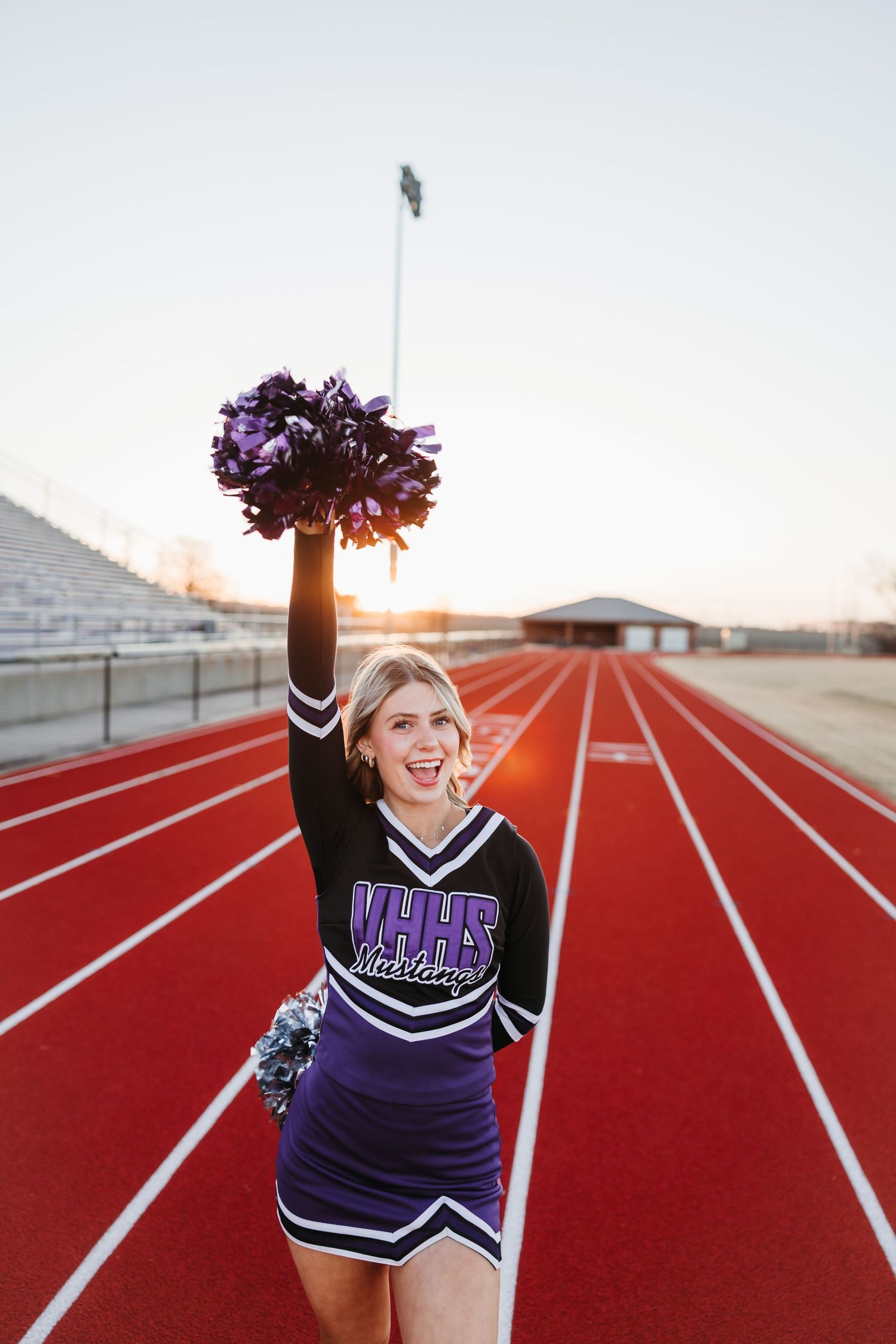 A cheerleader in a purple MYHS Mustangs uniform raises a pom-pom on a red track at sunset.