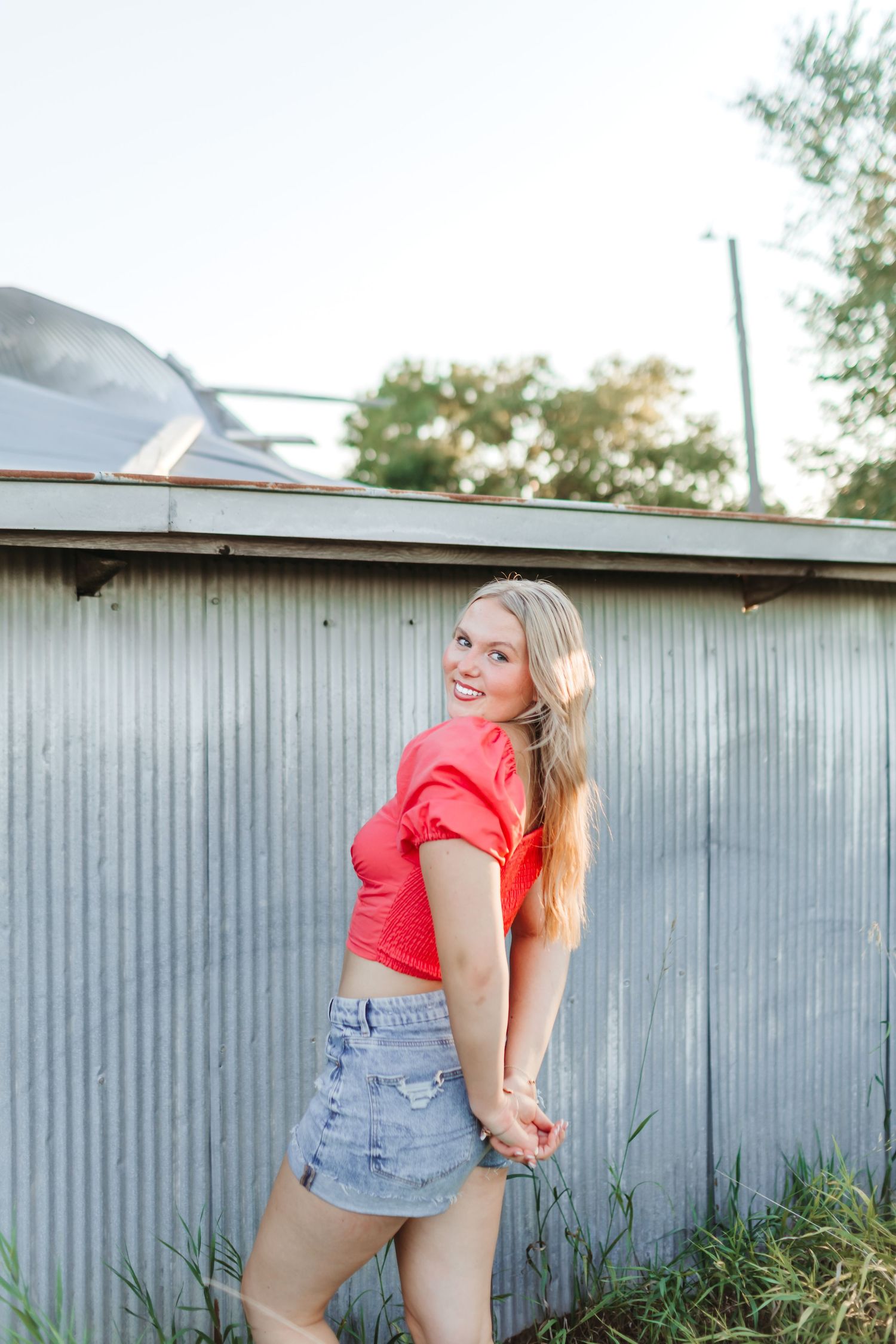 Smiling blonde woman in red crop top and denim mini skirt posing outdoors near corrugated metal wall.