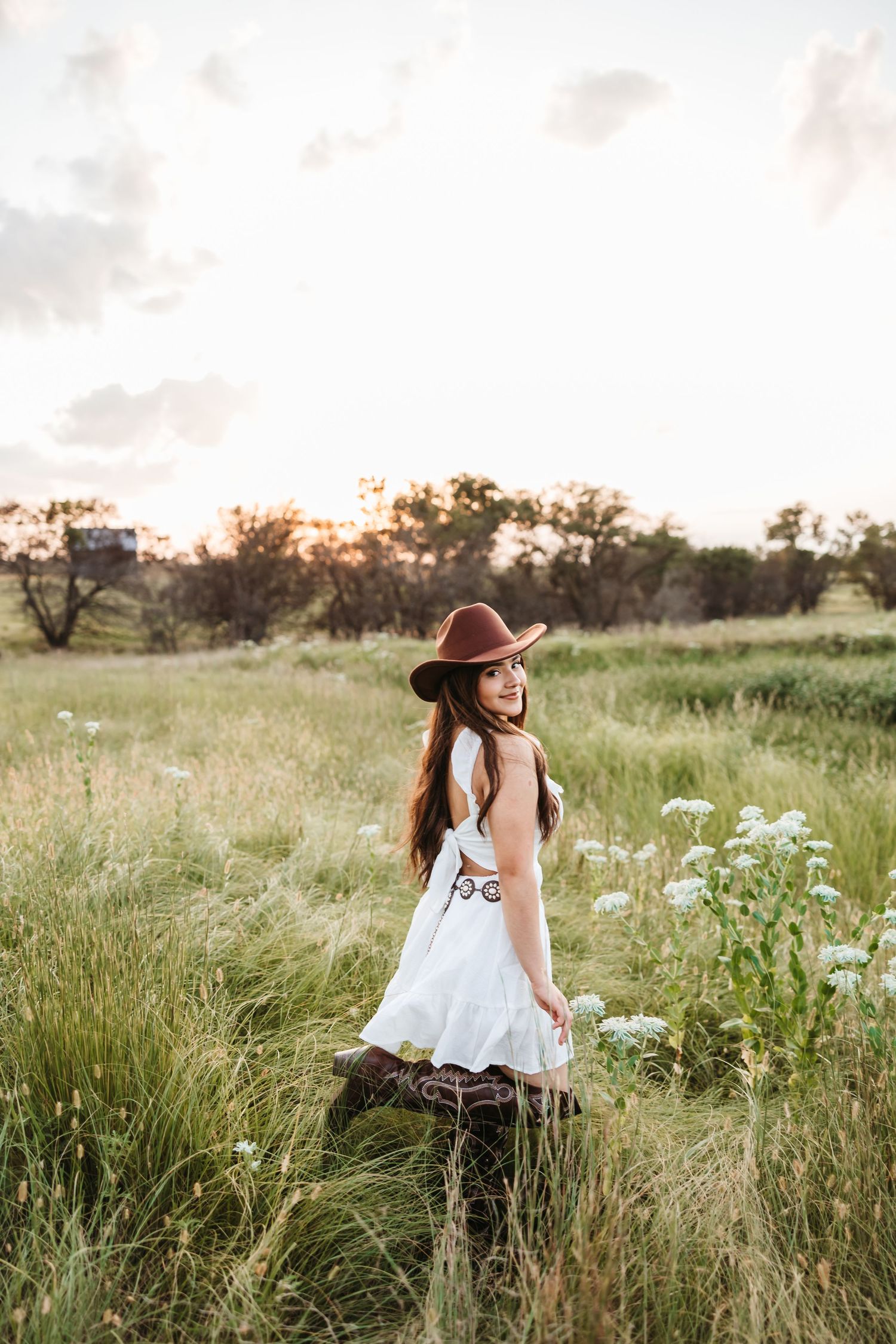 Person in white dress and hat walking through wildflower meadow at sunset.