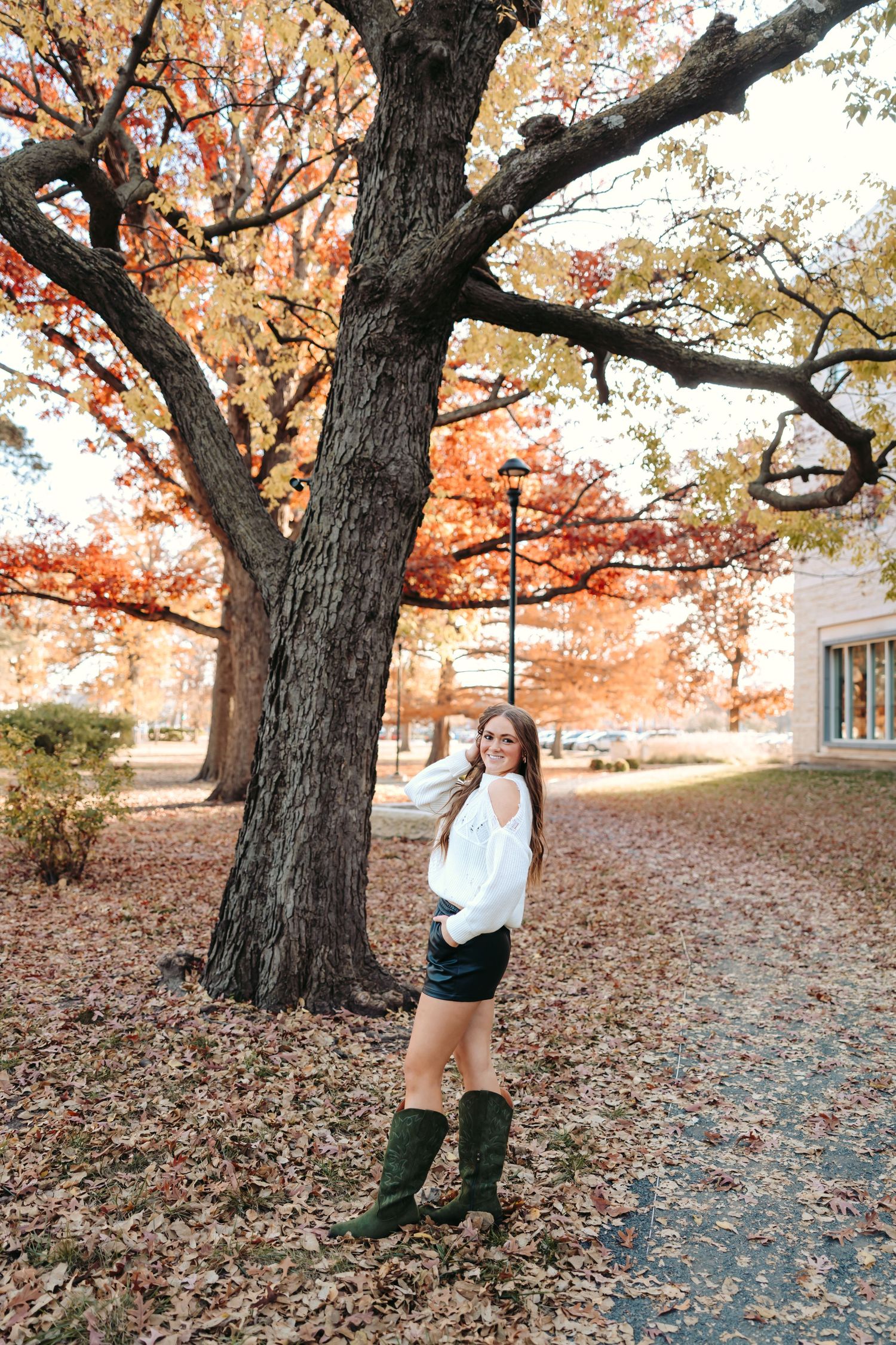 Woman beneath large autumn tree with orange leaves and fallen foliage.