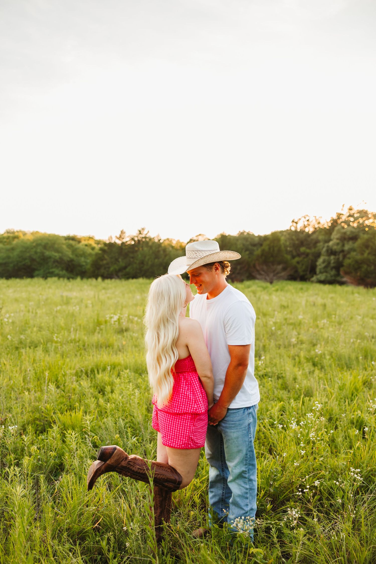 A couple in western attire embraces in a lush green field, woman in pink dress and boots, man in cowboy hat and jeans.