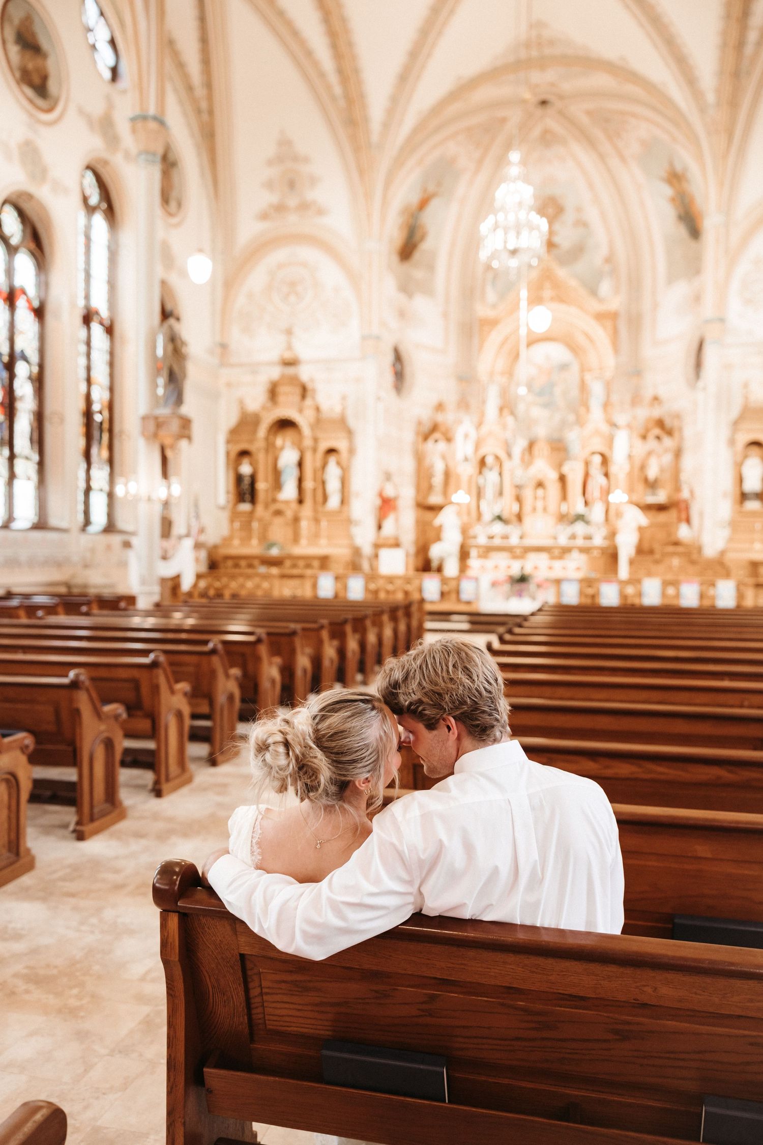 Couple in white sitting on church pew, sharing intimate moment before ornate altar in beautiful cathedral.