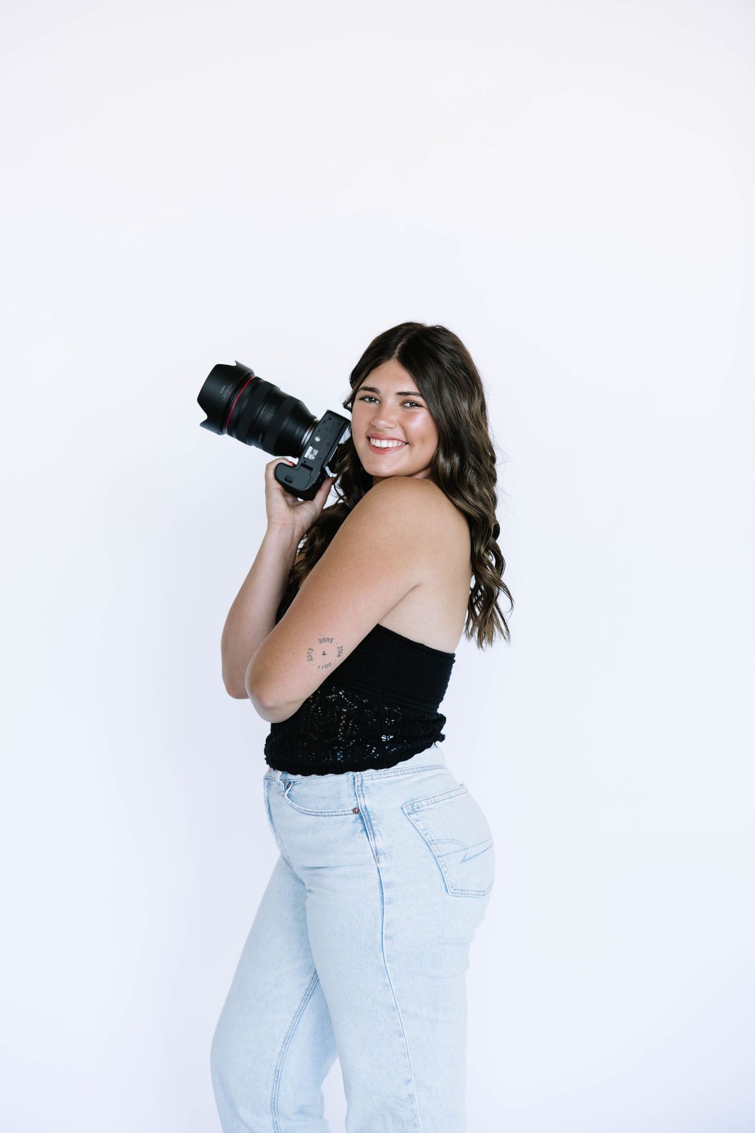 Professional photographer in black top and jeans posing with camera against white wall.