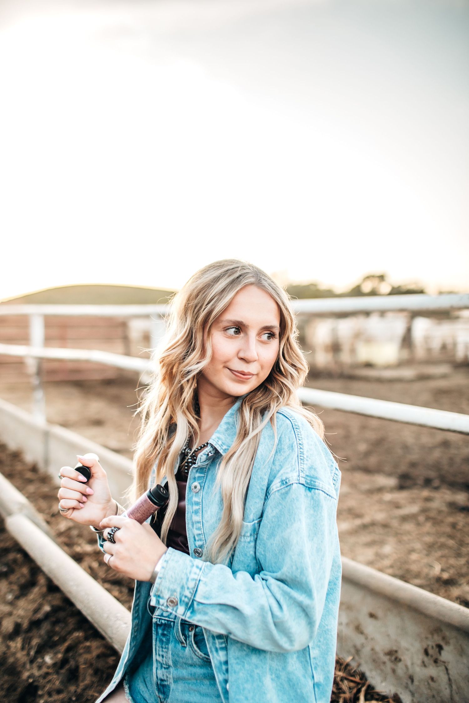 Woman in light blue denim jacket poses near white fence at sunset.