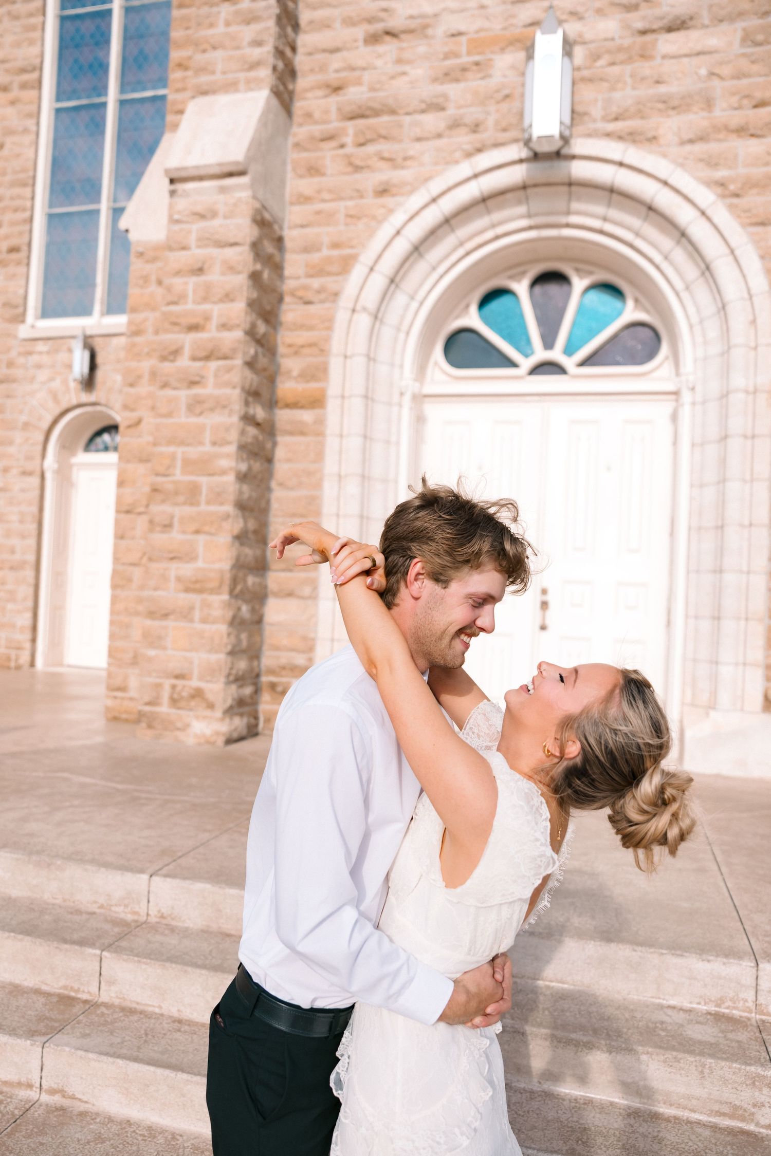 Bride and groom embracing and laughing together in front of a stone church with stained glass windows on their wedding day.