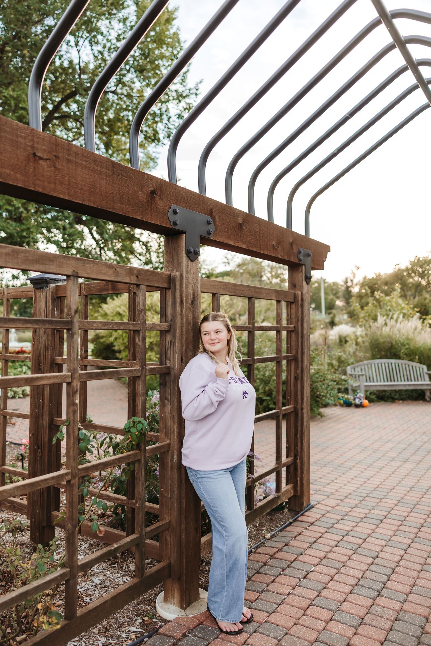 Young woman in lavender sweater and jeans posing by a wooden pergola in a garden setting.