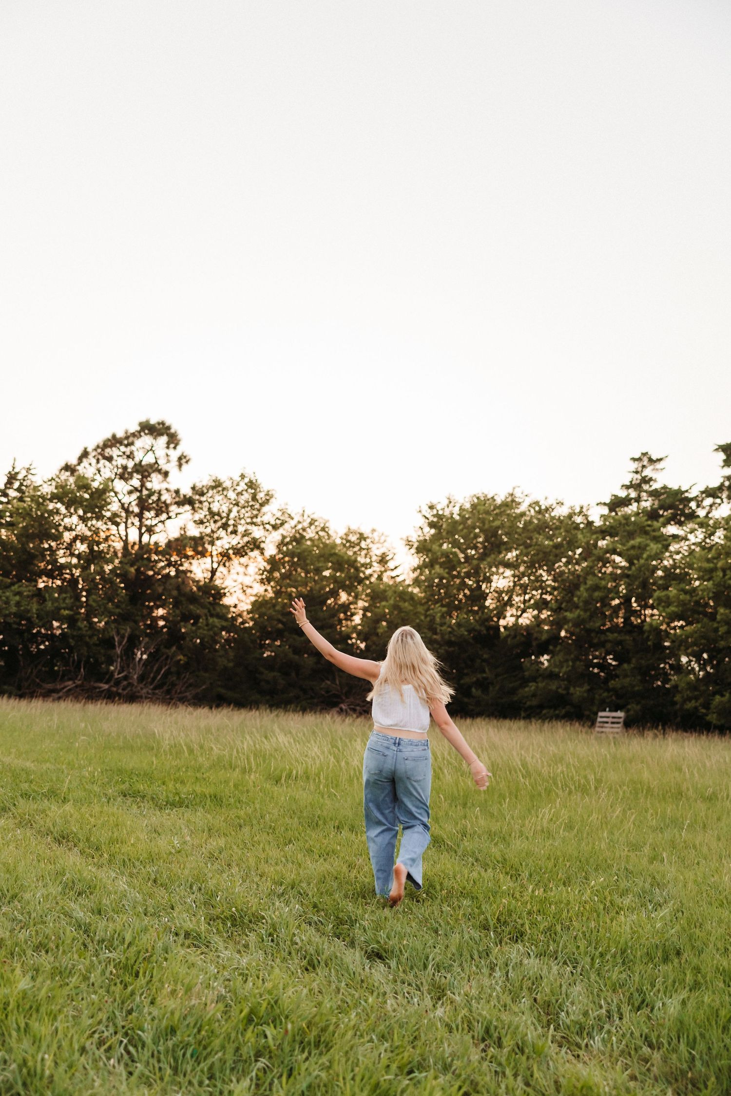 Blonde woman in white top and jeans walking through a green field at sunset with trees in background.