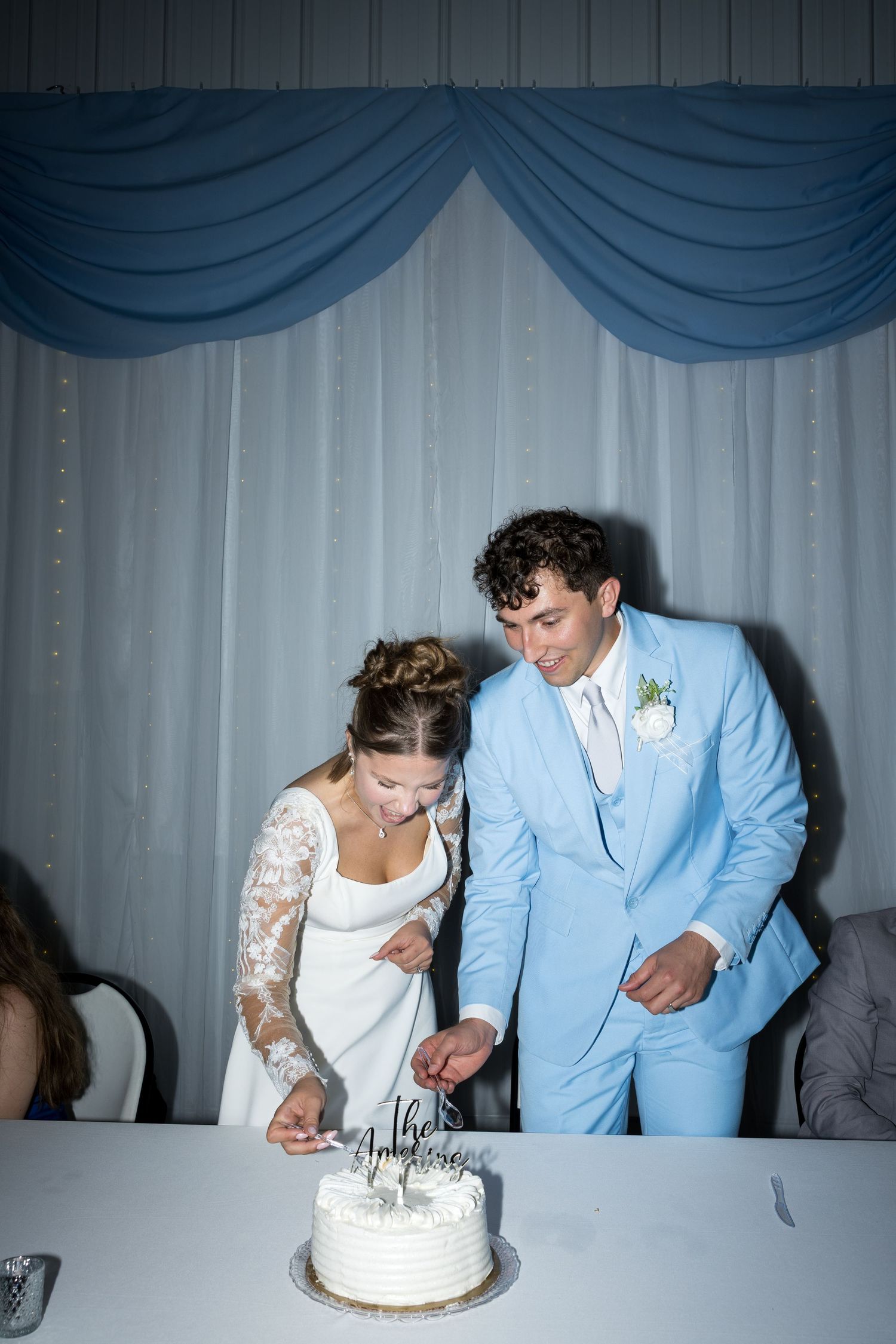 Bride and groom in light blue suit cut white wedding cake together at elegant blue and white draped reception venue.