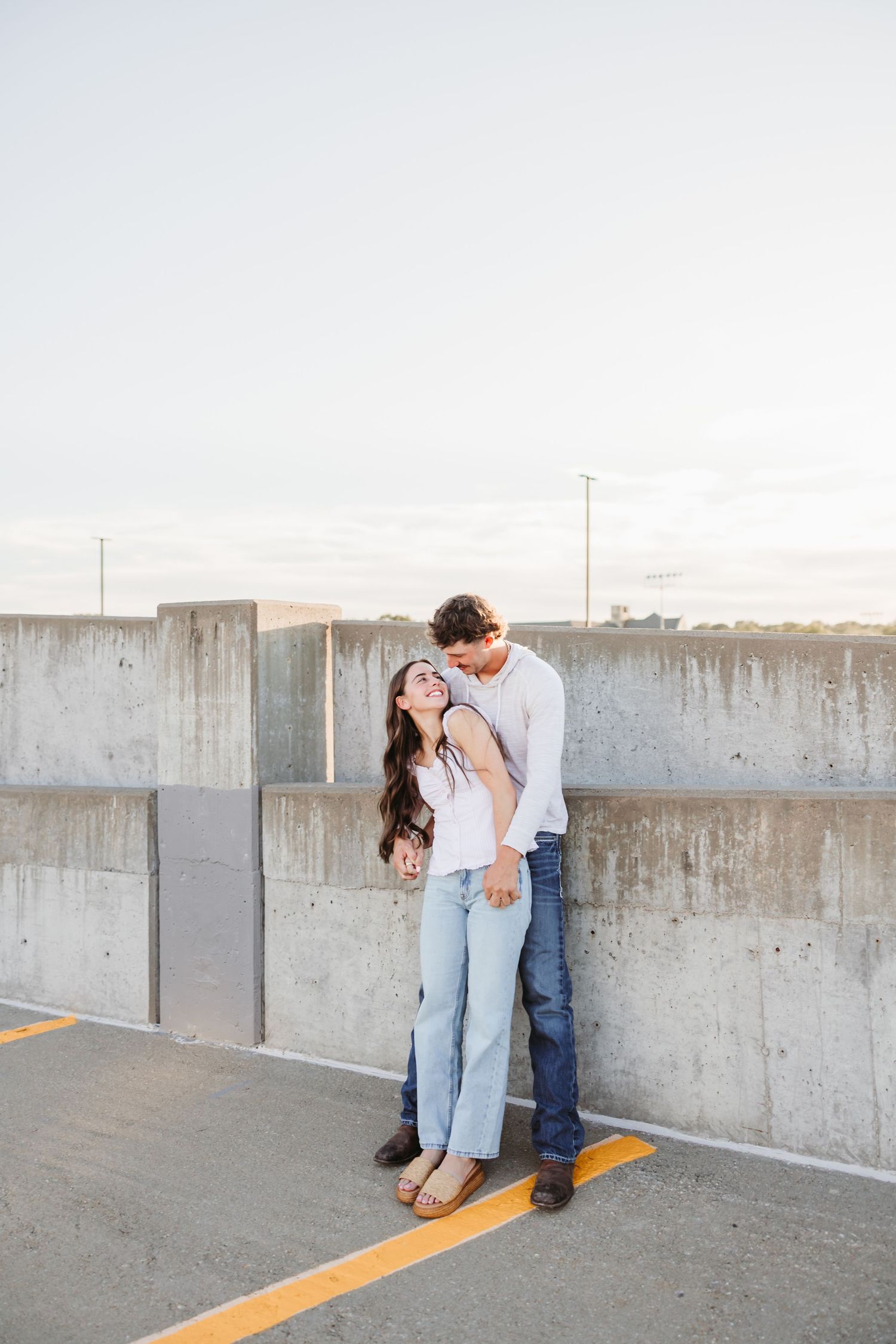 Couple poses against concrete wall at the top of a parking garage.
