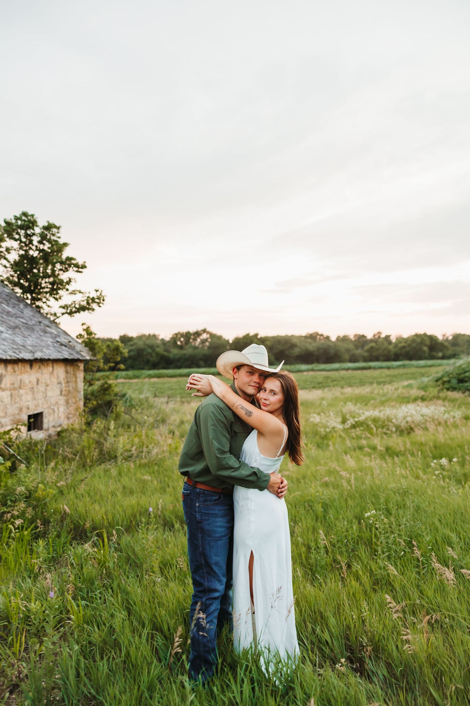 Couple embracing in a lush green field at sunset, man in cowboy hat and green shirt, woman in white dress.