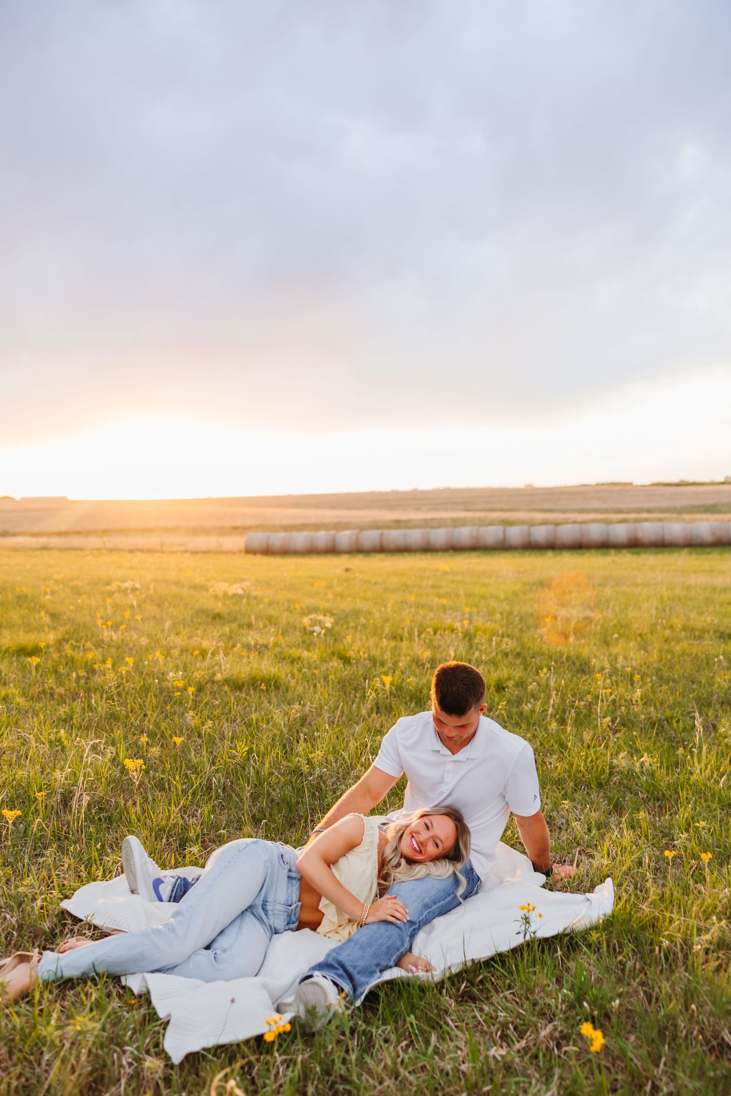 Couple relaxing on a blanket in a sunny green field at sunset, with hay bales in the background.