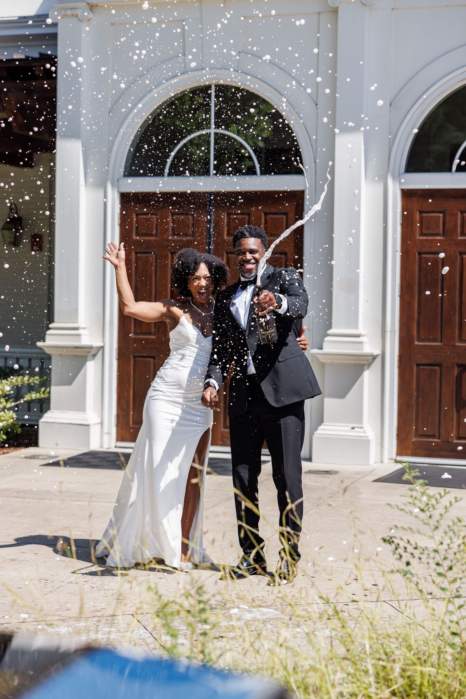 Joyful newlywed couple celebrating outside a white church with confetti, bride in white gown and groom in black suit.