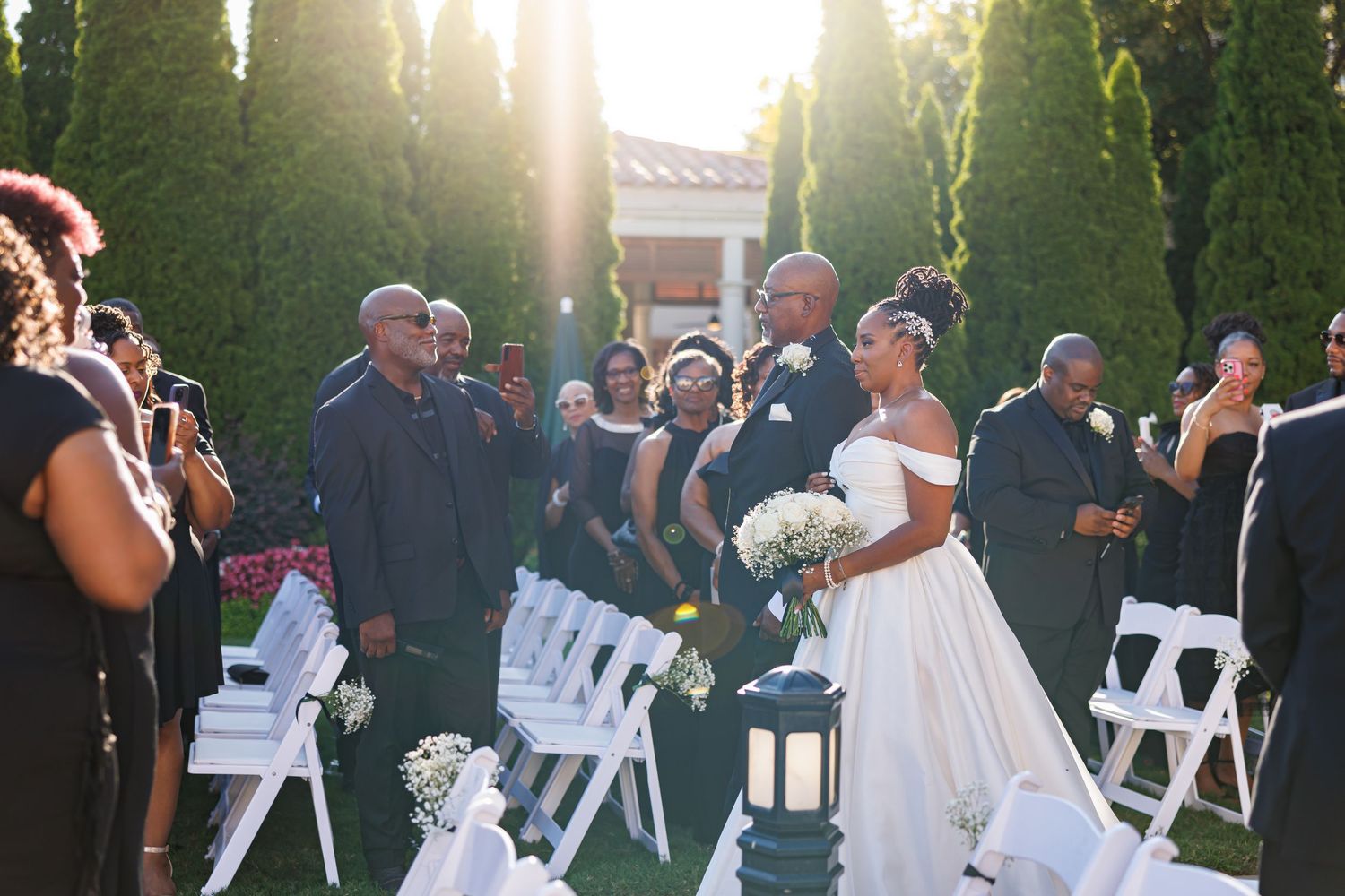 Luxury wedding picture of Bride in off-shoulder gown walking down outdoor aisle with escort at sunset wedding ceremony with cypress trees.