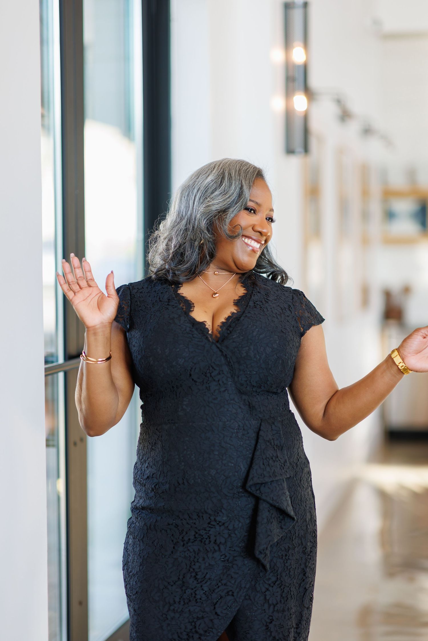High-end picture of Joyful older Black woman with gray hair laughs in black lace dress against bright window wall.