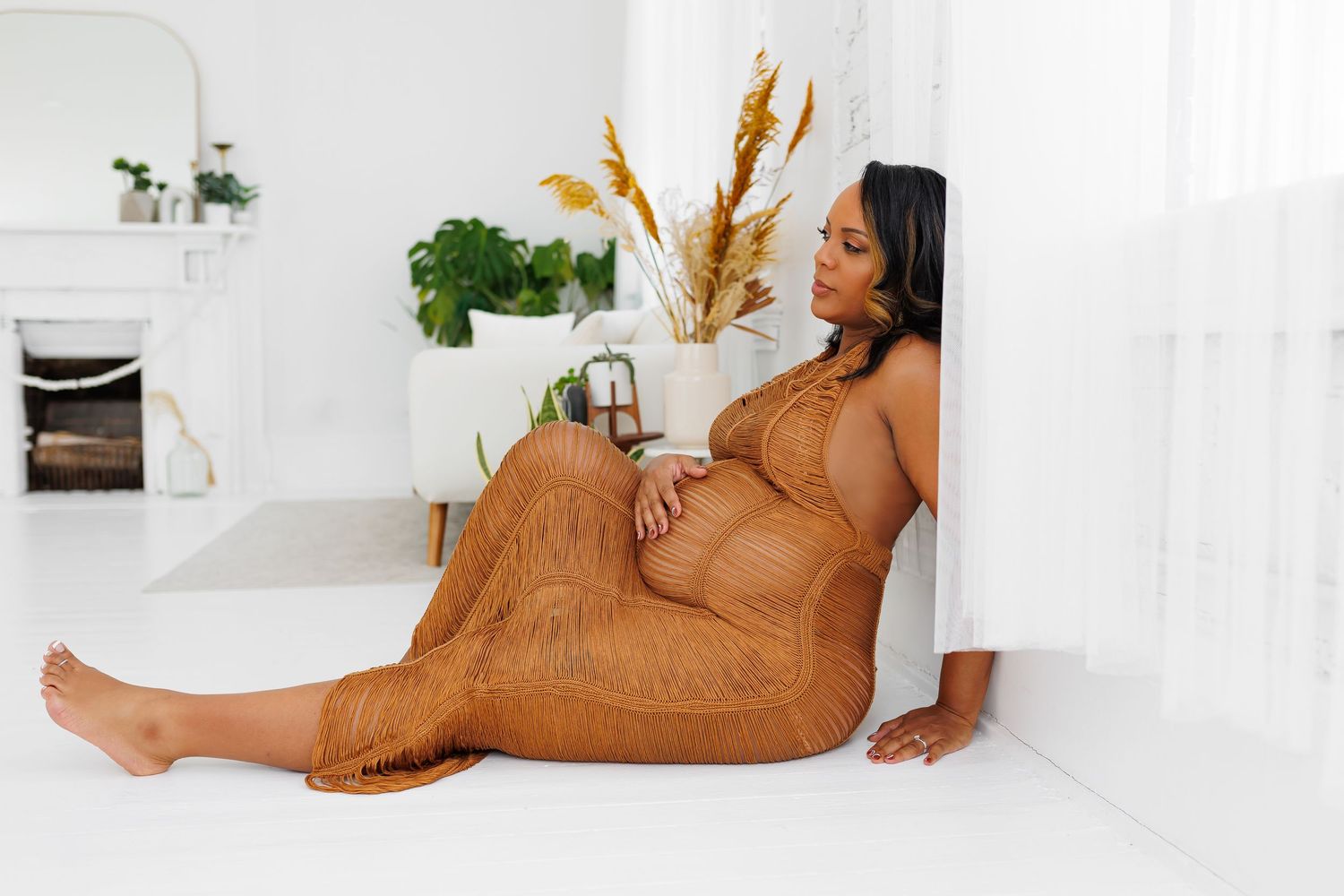Luxury picture of A pregnant woman in a brown lace dress sits on a white floor leaning against a wall in a bright room.