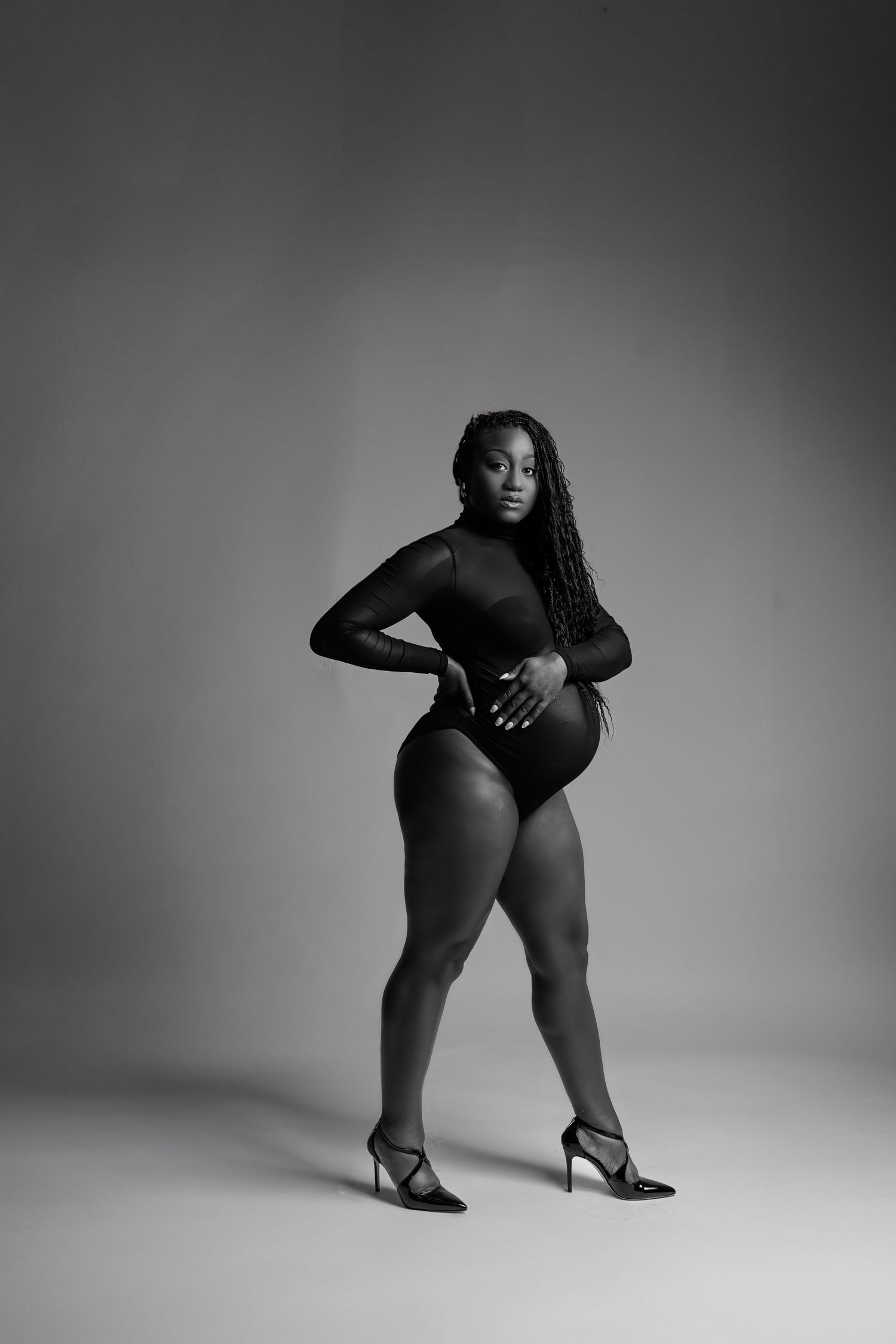 Black and white studio portrait of a confident woman in bodysuit and heels posing gracefully.