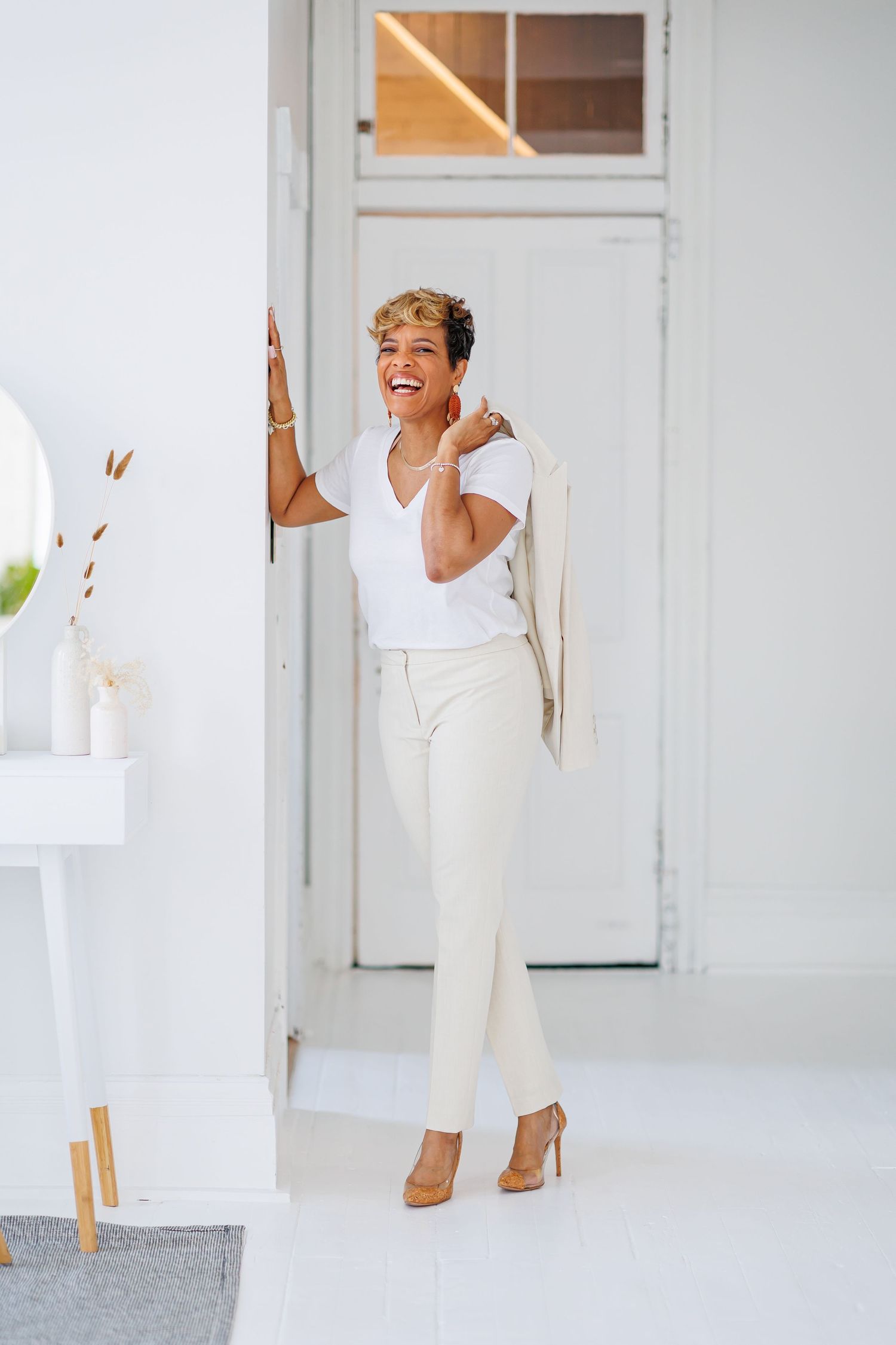 Natural light picture of Stylish woman in white outfit laughing while leaning against doorframe in bright white room.