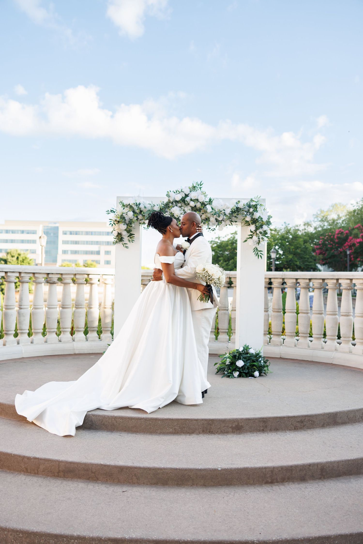 Luxury pictures of Bride and groom sharing romantic kiss on elegant terrace with white balustrade and floral wedding arch.