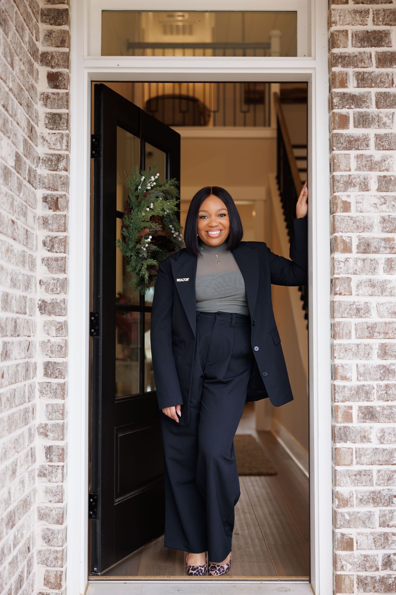 Timeless picture of Smiling woman in black blazer and wide-leg pants standing in doorway of brick home.