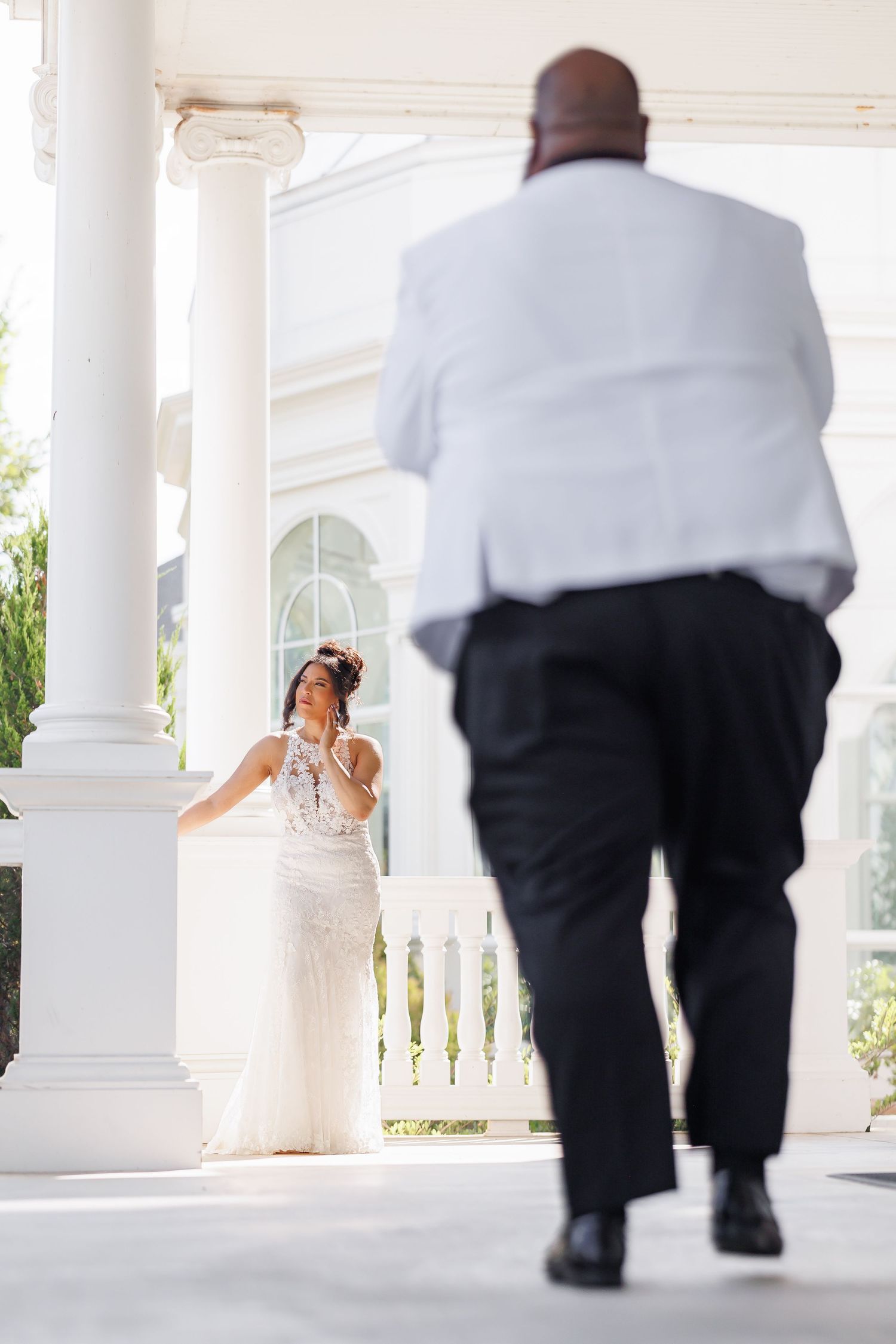 Elegant picture of Bride in lace gown awaits groom approaching columned venue entrance for first look moment.