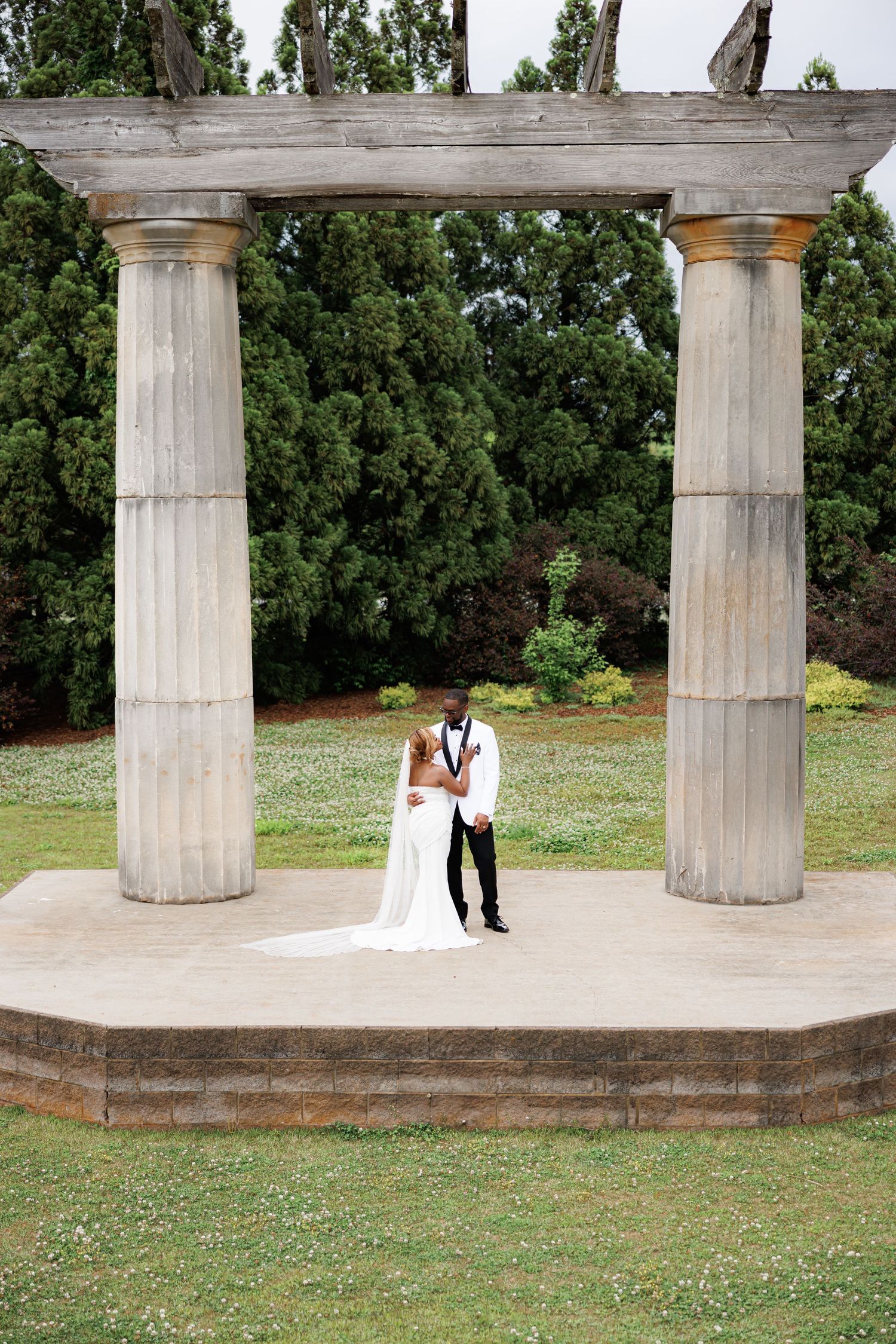 Luxury wedding picture of Bride and groom in white wedding attire stand together beneath elegant stone columns in a botanical gardens.