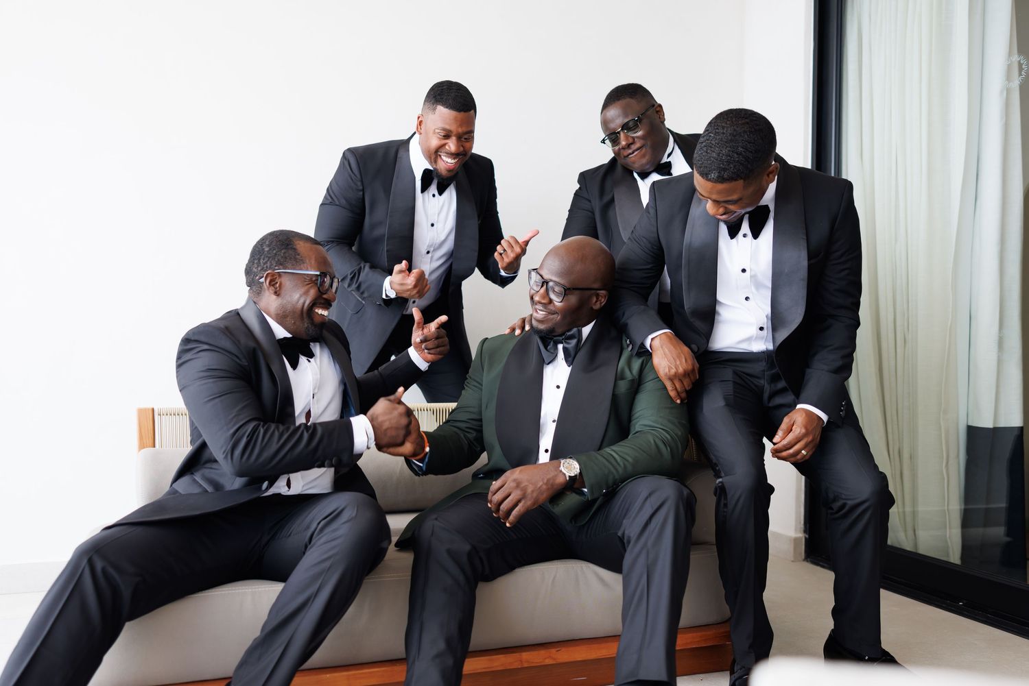 Elegant picture of Five groomsmen in black tuxedos laugh together on a sofa before the wedding ceremony.