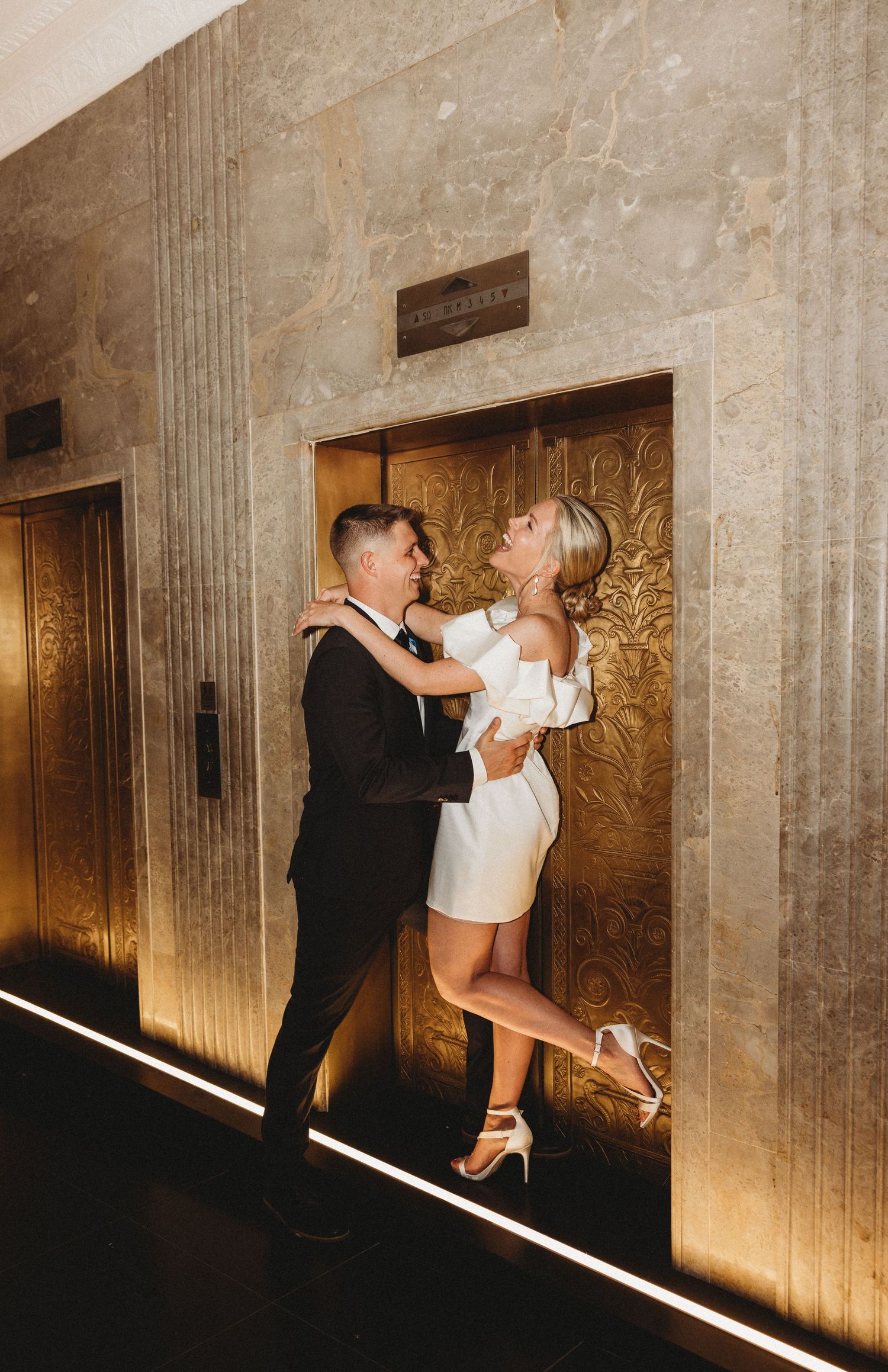 Couple embracing and laughing in front of ornate gold elevator doors, woman in white mini dress and heels, man in black suit.