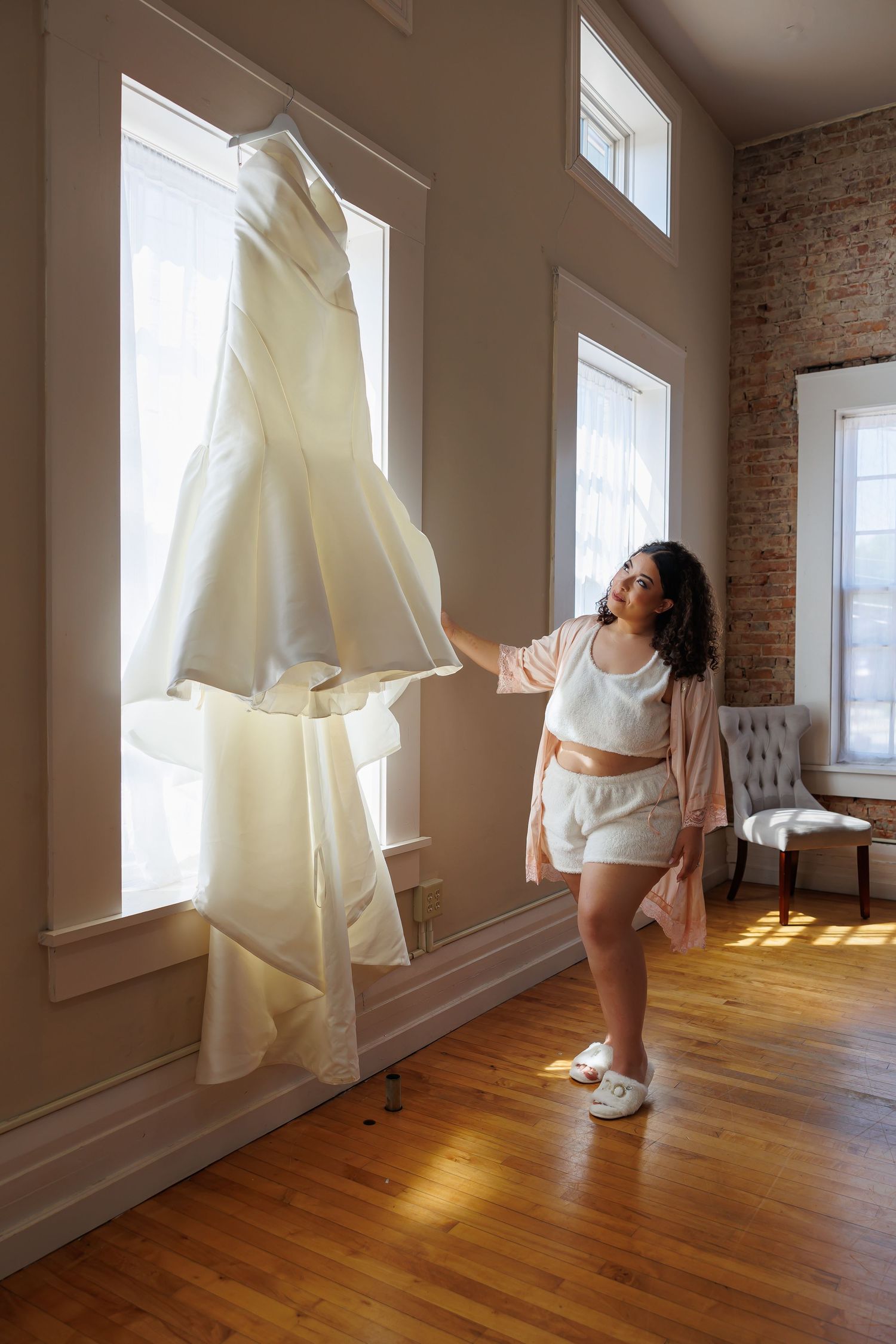 Luxury wedding picture ofBride in robe admires her white wedding gown hanging in a sunlit room with tall windows.