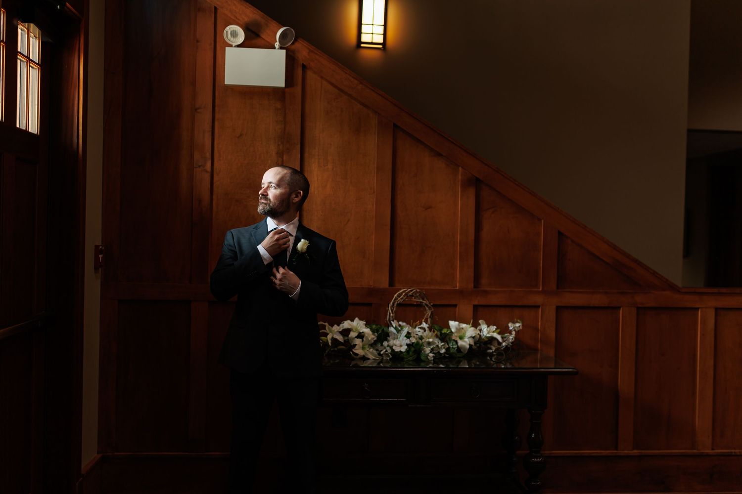 Luxury wedding picture of Groom adjusts his tie in dramatic lighting beside a floral arrangement in a wood-paneled hall.