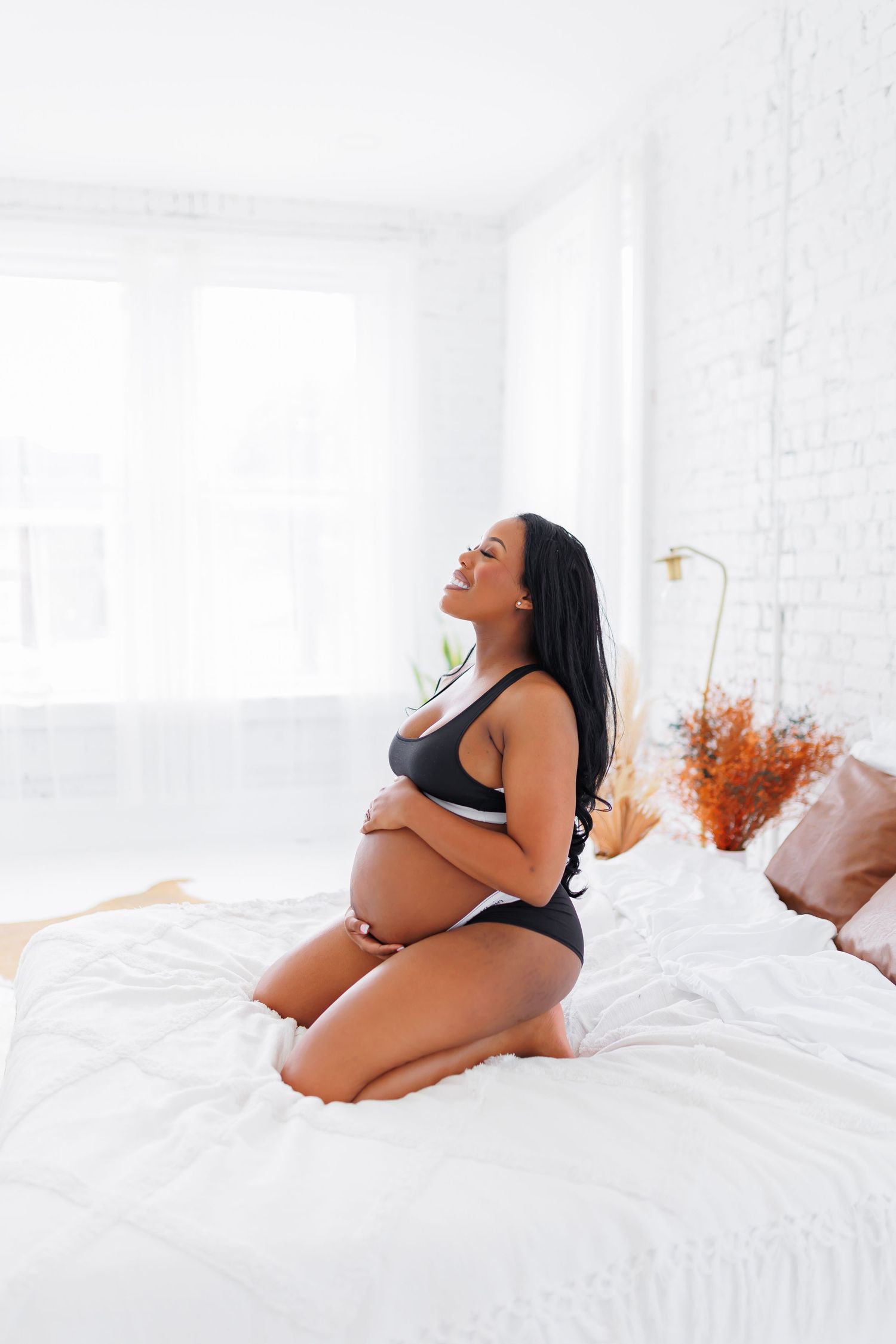 Natural light studio portrait of A pregnant woman in black lingerie kneels on a white bed with eyes closed, hands on her belly.