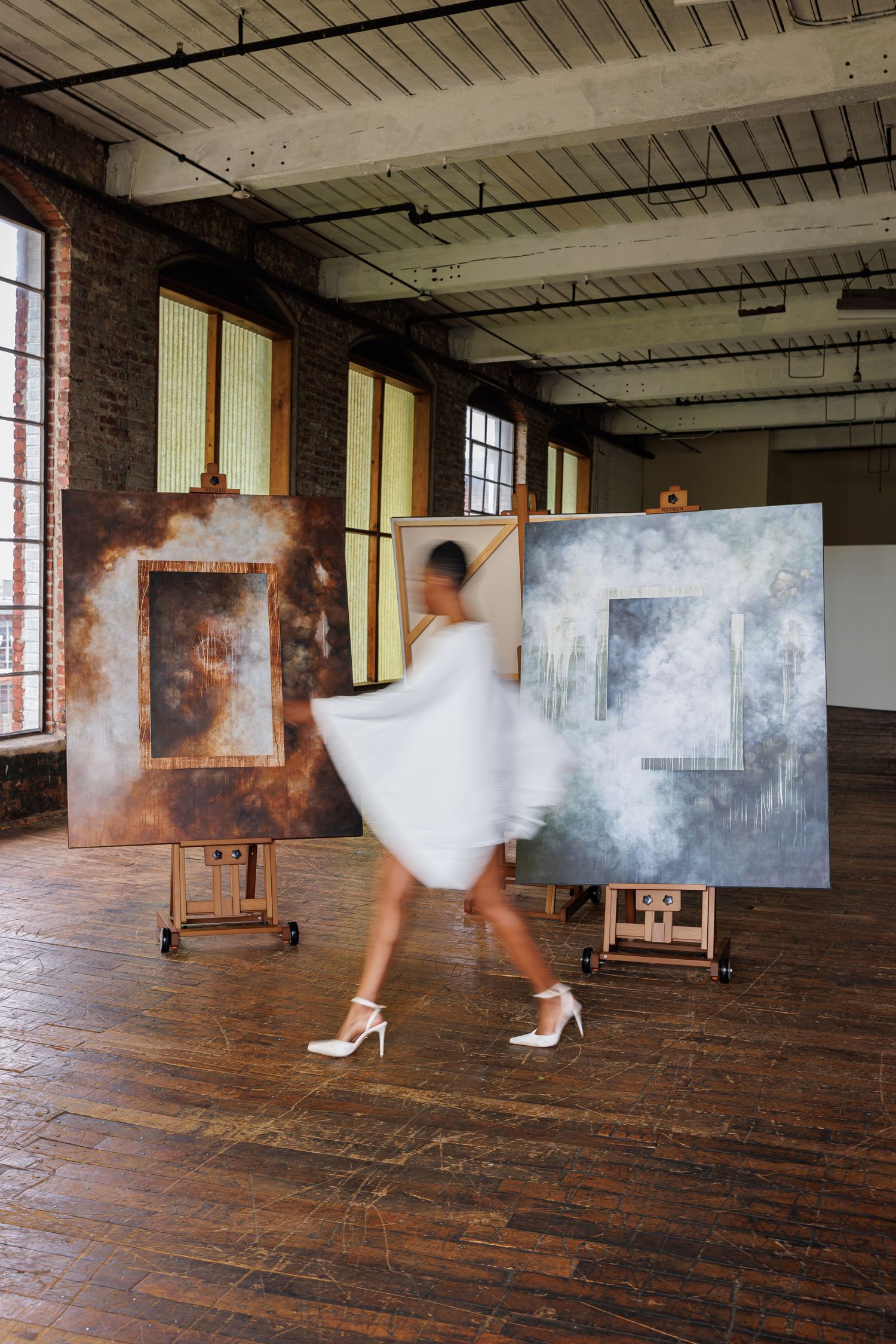 Blurred figure in white dress walking past large paintings on easels in a spacious brick-walled art studio.
