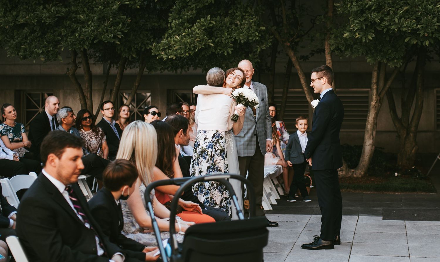 Wedding ceremony guests embrace while groom waits at the altar during an outdoor celebration.