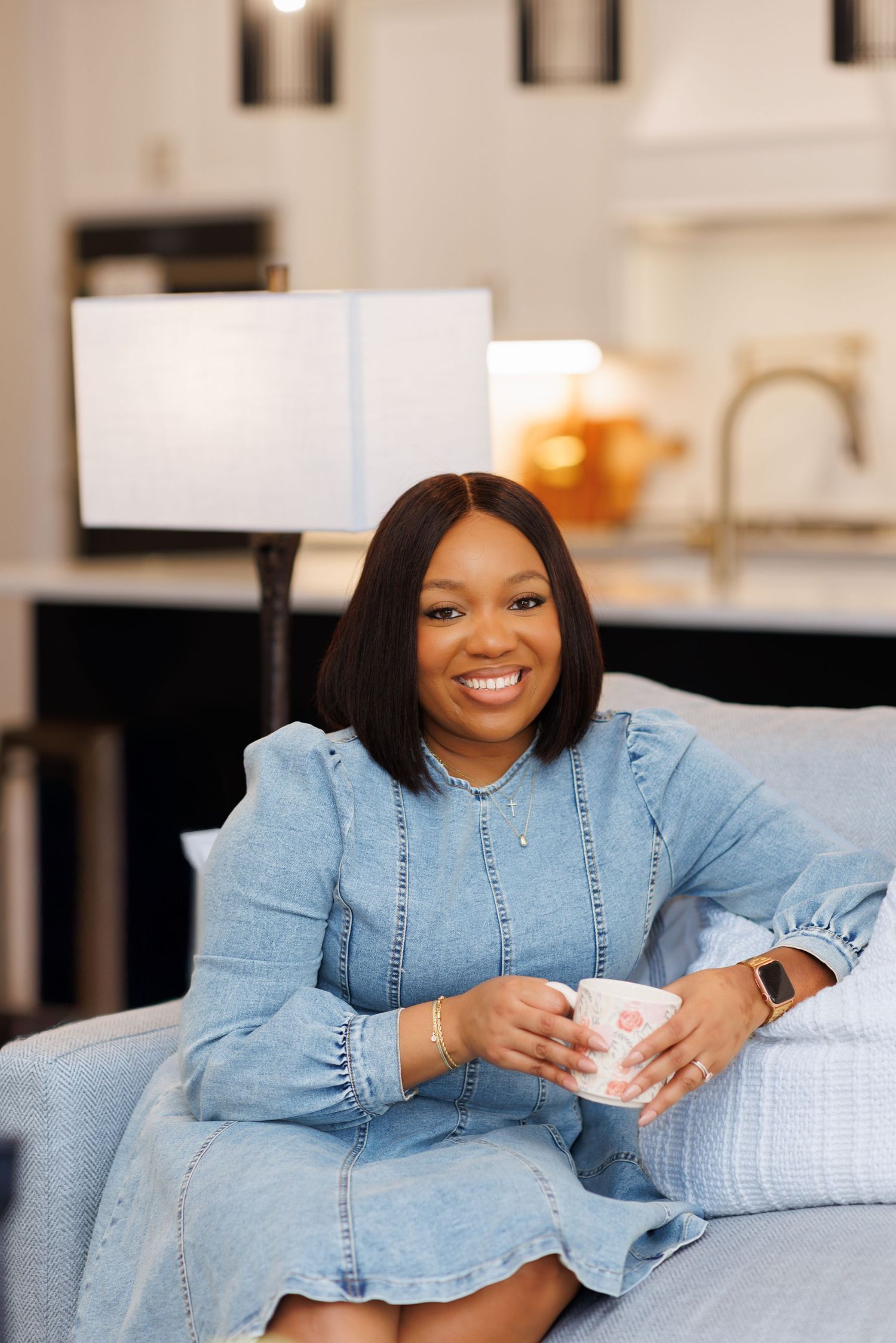 Elegant picture of Woman in denim dress sitting on couch holding floral mug and smiling warmly.