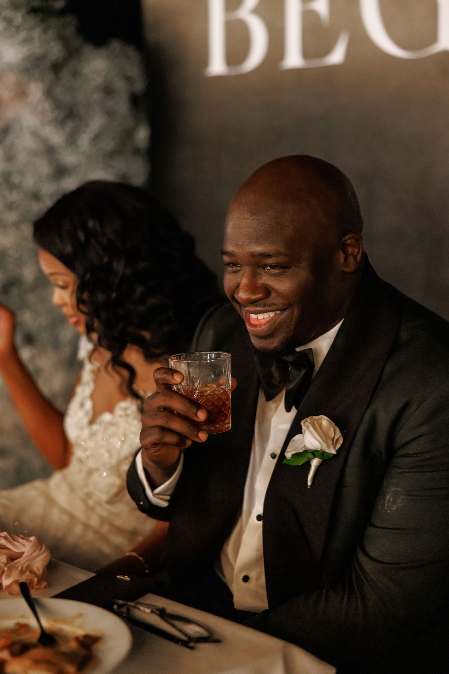 Timeless picture of Smiling groom in tuxedo holding a cocktail glass at wedding reception sweetheart table with bride beside him.