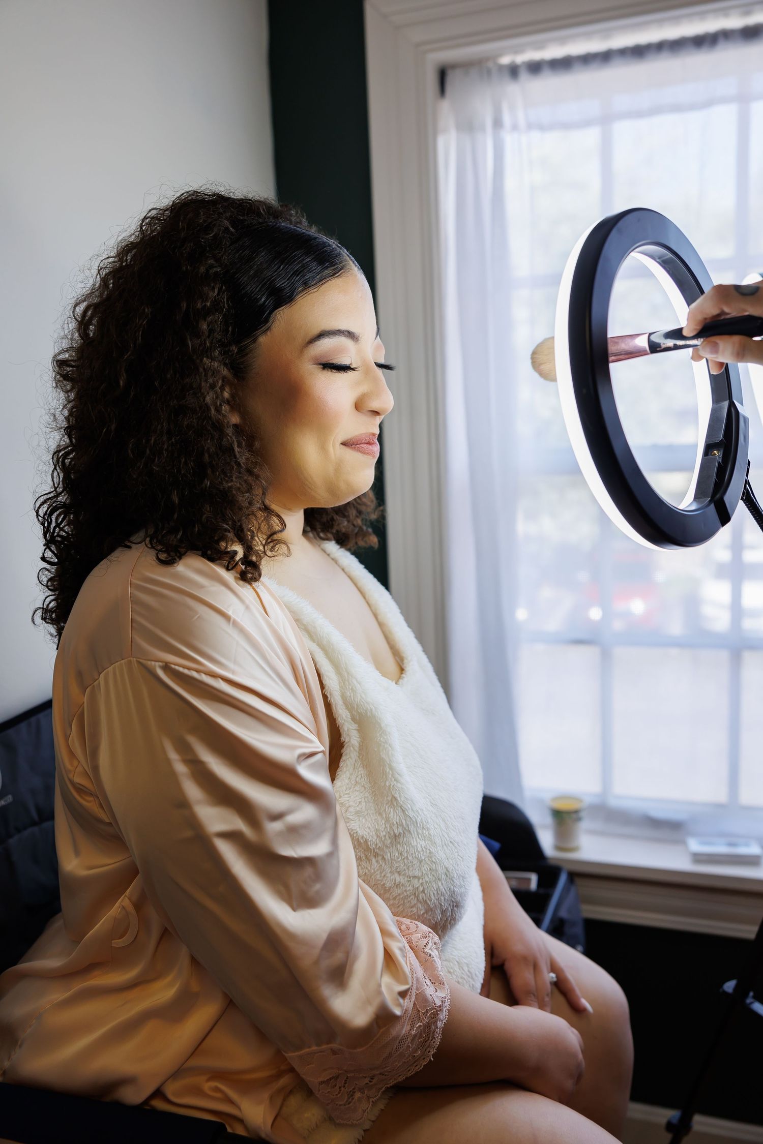 Luxury wedding picture of Bride smiles with eyes closed while makeup artist applies cosmetics using a ring light.