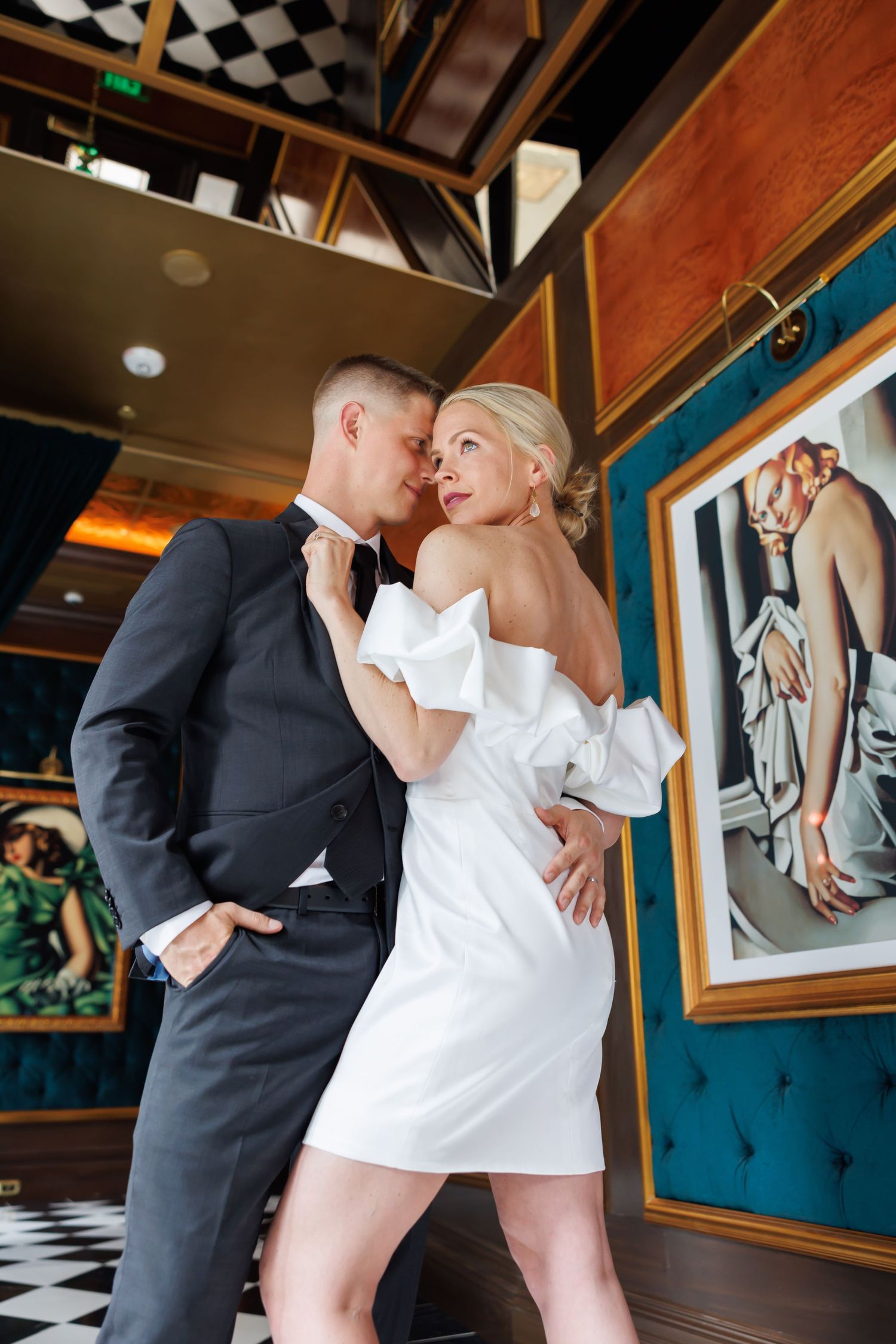 Elegant couple in formal attire posing romantically in a luxurious interior with framed artwork on teal walls.