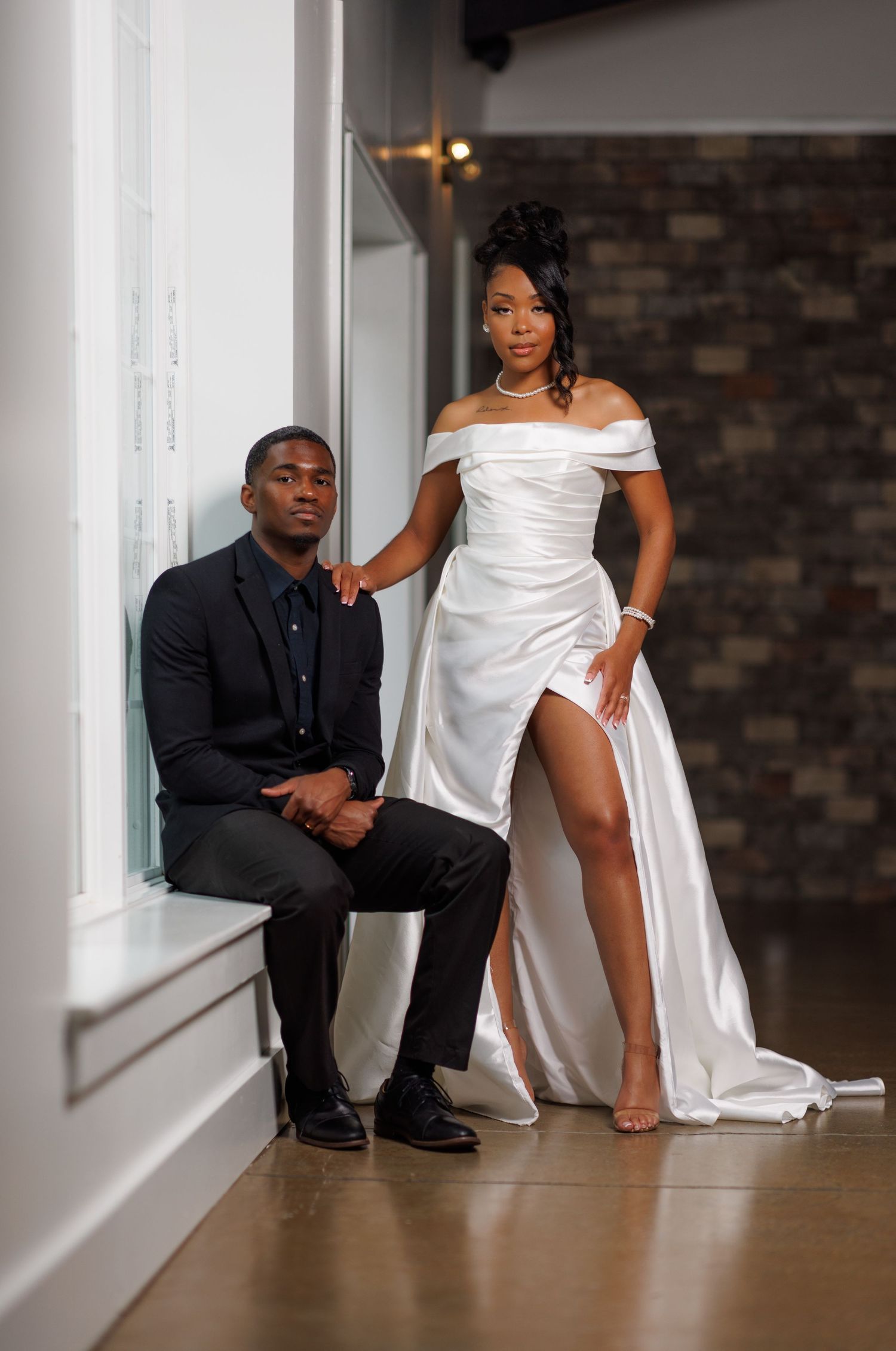 Luxury wedding picture of Bride in white satin gown stands beside groom in black suit by bright venue windows.