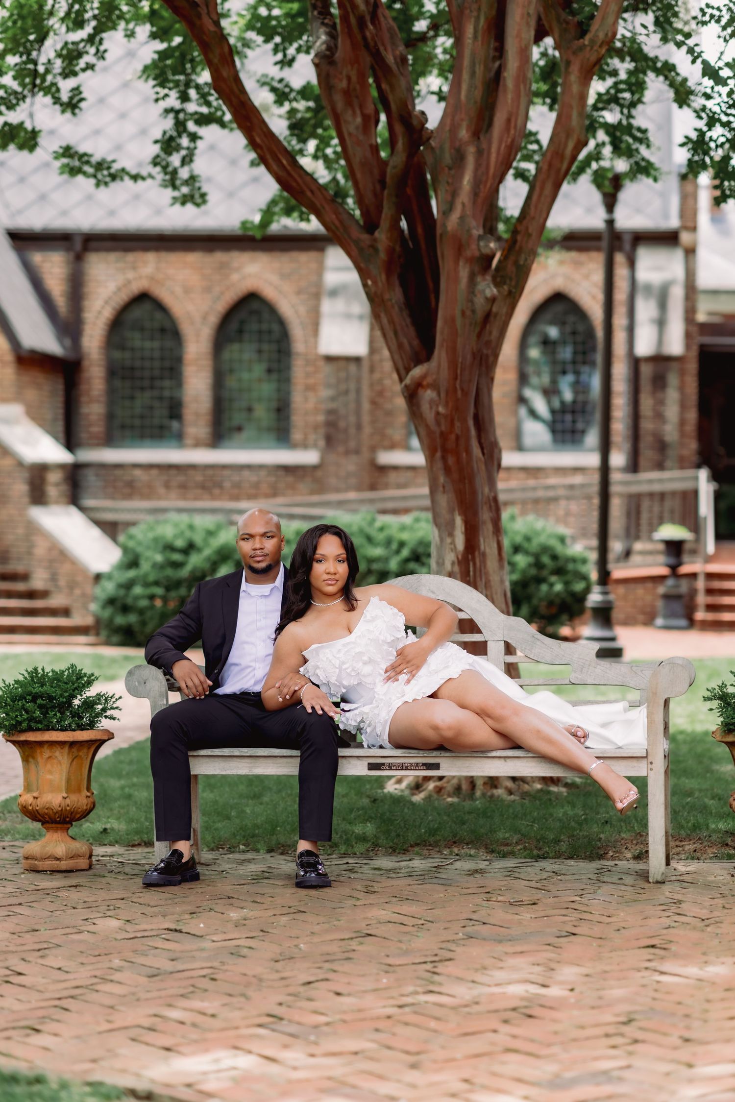 Elegant couple posing on a white bench outside a brick church, woman in white dress, man in dark suit.