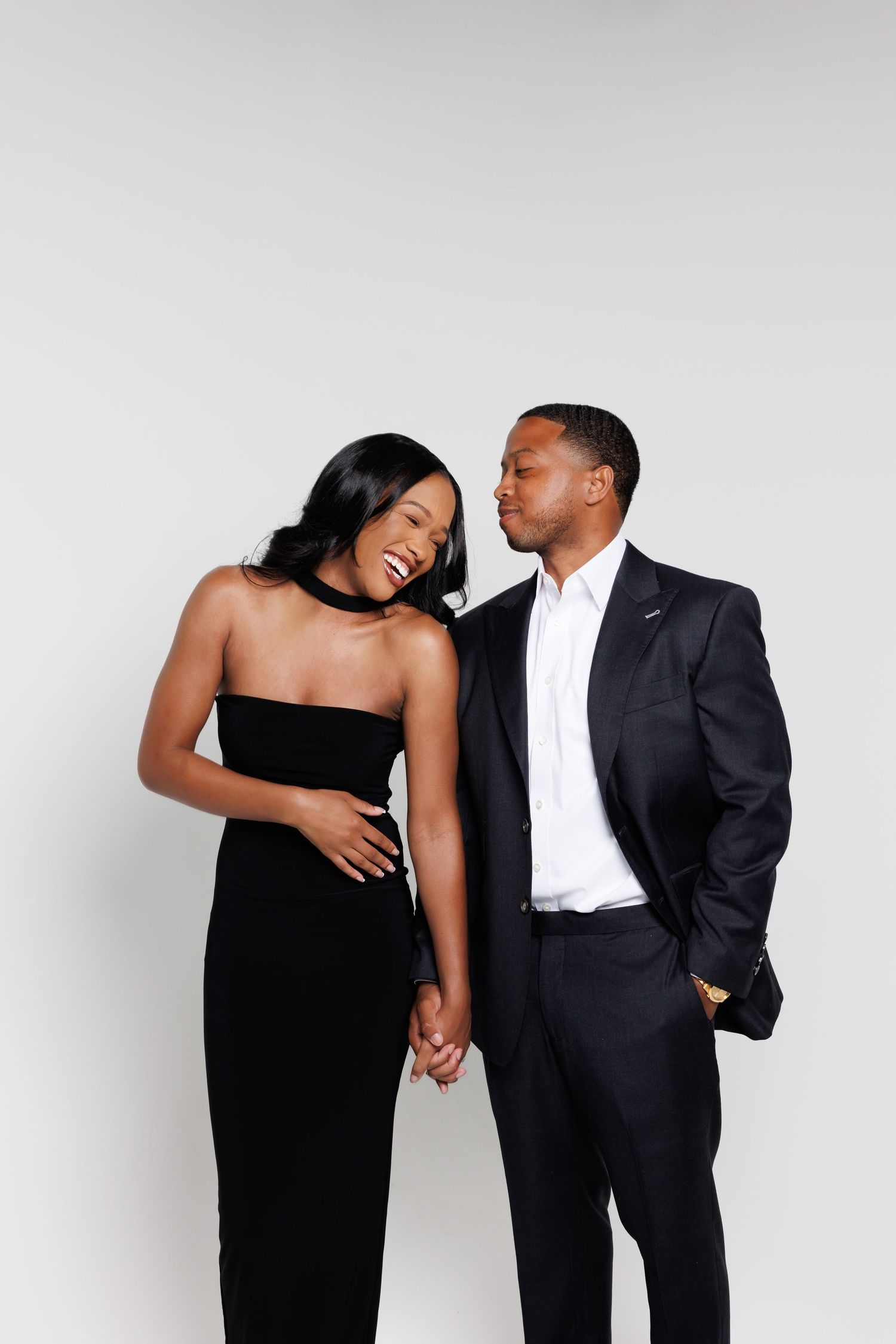 Elegant couple in matching black formal attire smiling together against a white background.