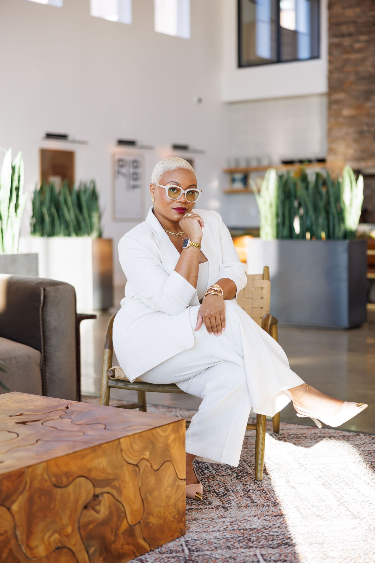Luxury picture of Stylish Black woman in white pantsuit and cat-eye sunglasses seated in modern lobby.