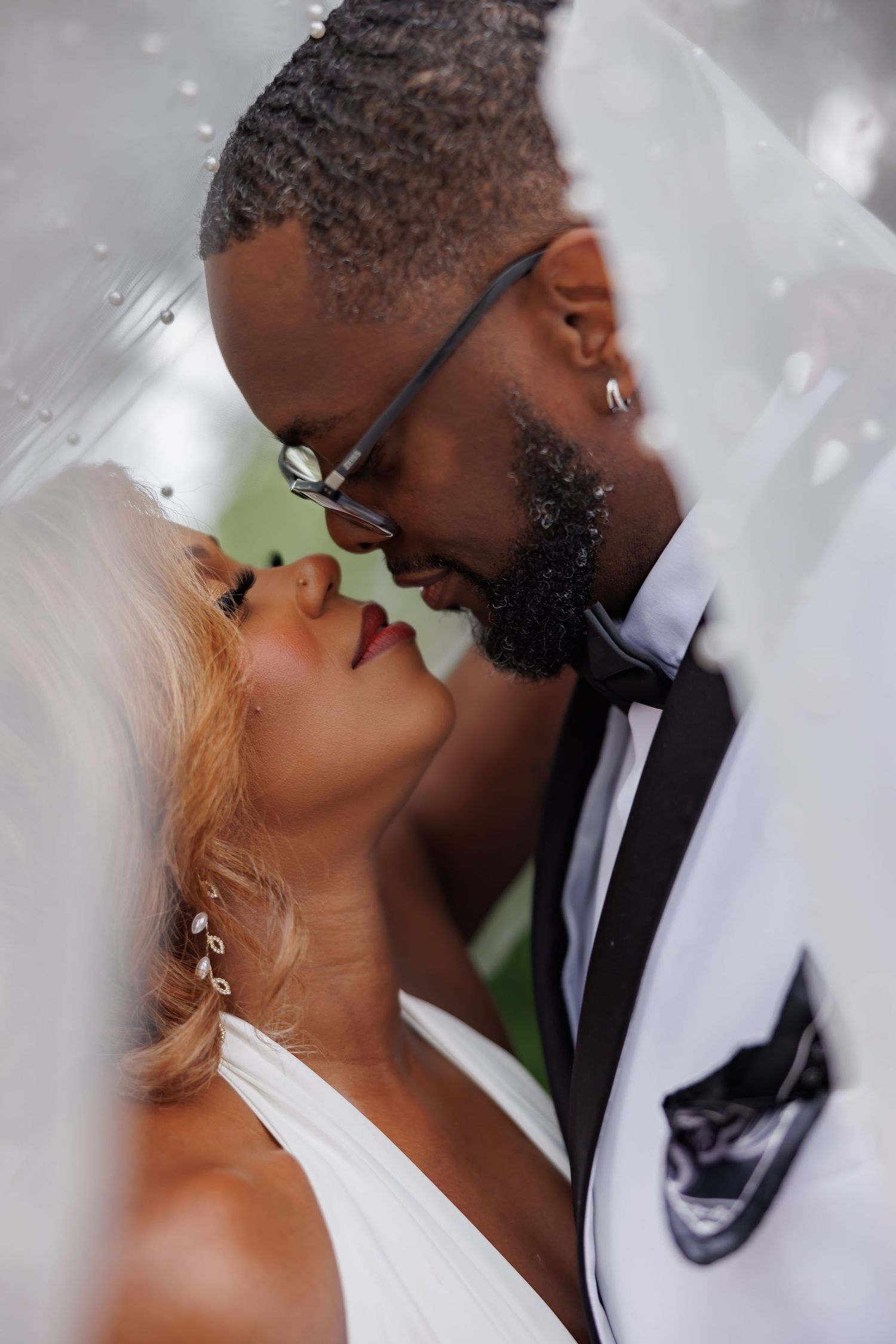 A bride and groom share a romantic almost-kiss under a wedding veil, dressed in bridal white and a tuxedo.