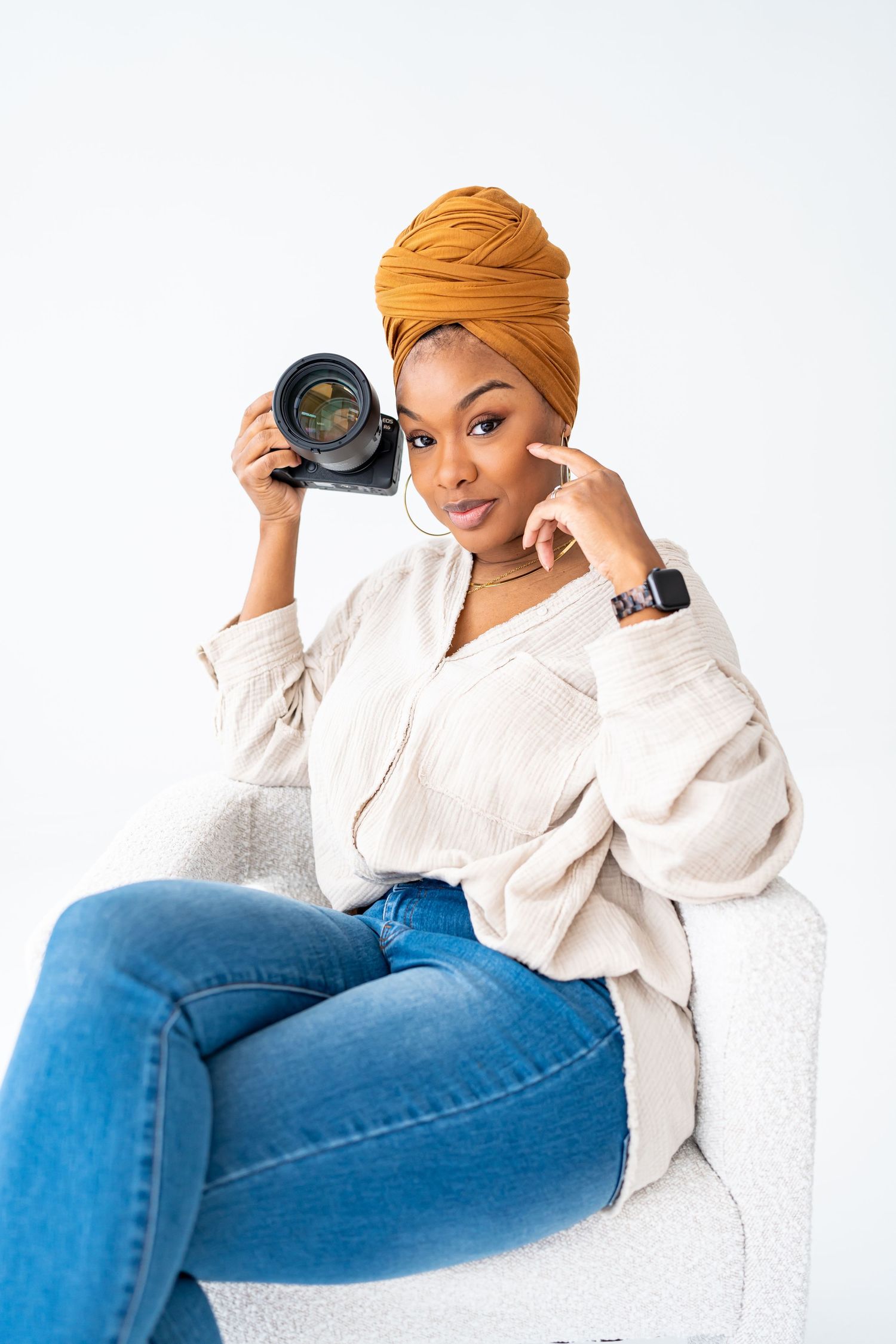 Natural light picture of Black female photographer poses confidently in a white blouse and jeans, holding a camera while seated in a chair.