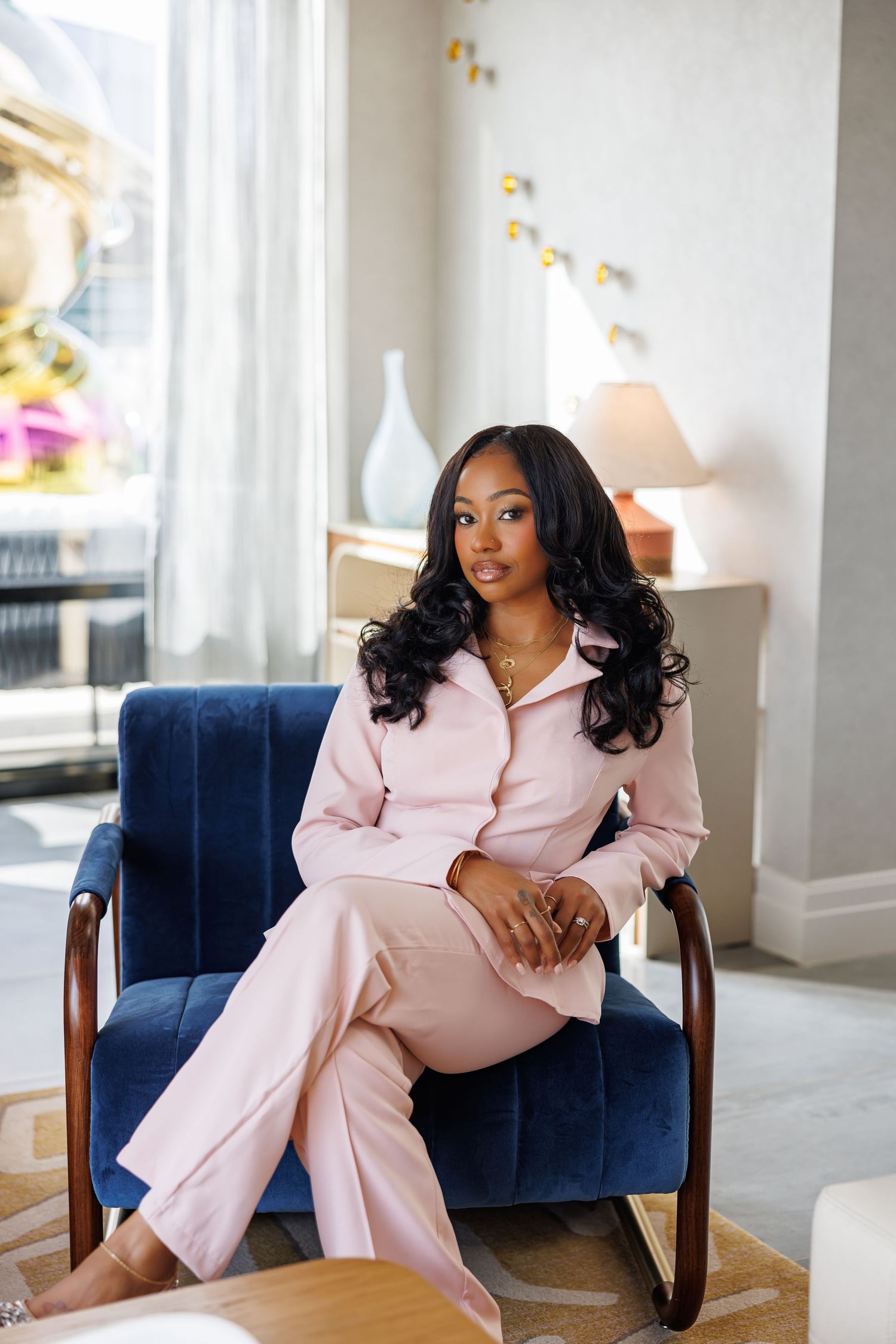 Elegant picture of young Black woman in pink pantsuit sits relaxed in navy velvet armchair by window.