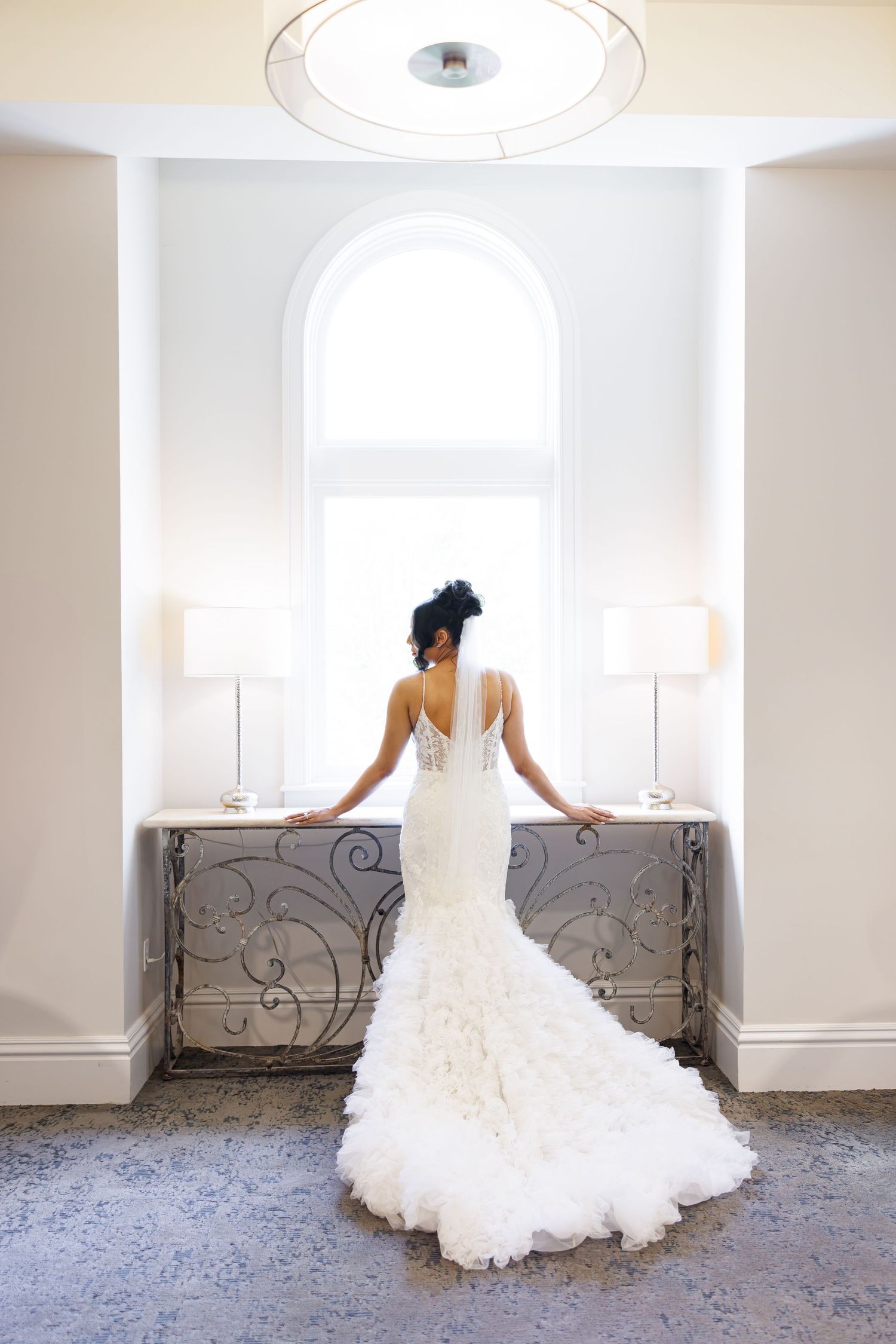 Luxury wedding picture of Elegant bride in backless mermaid wedding gown with ruffled train standing before arched window in luxury venue.
