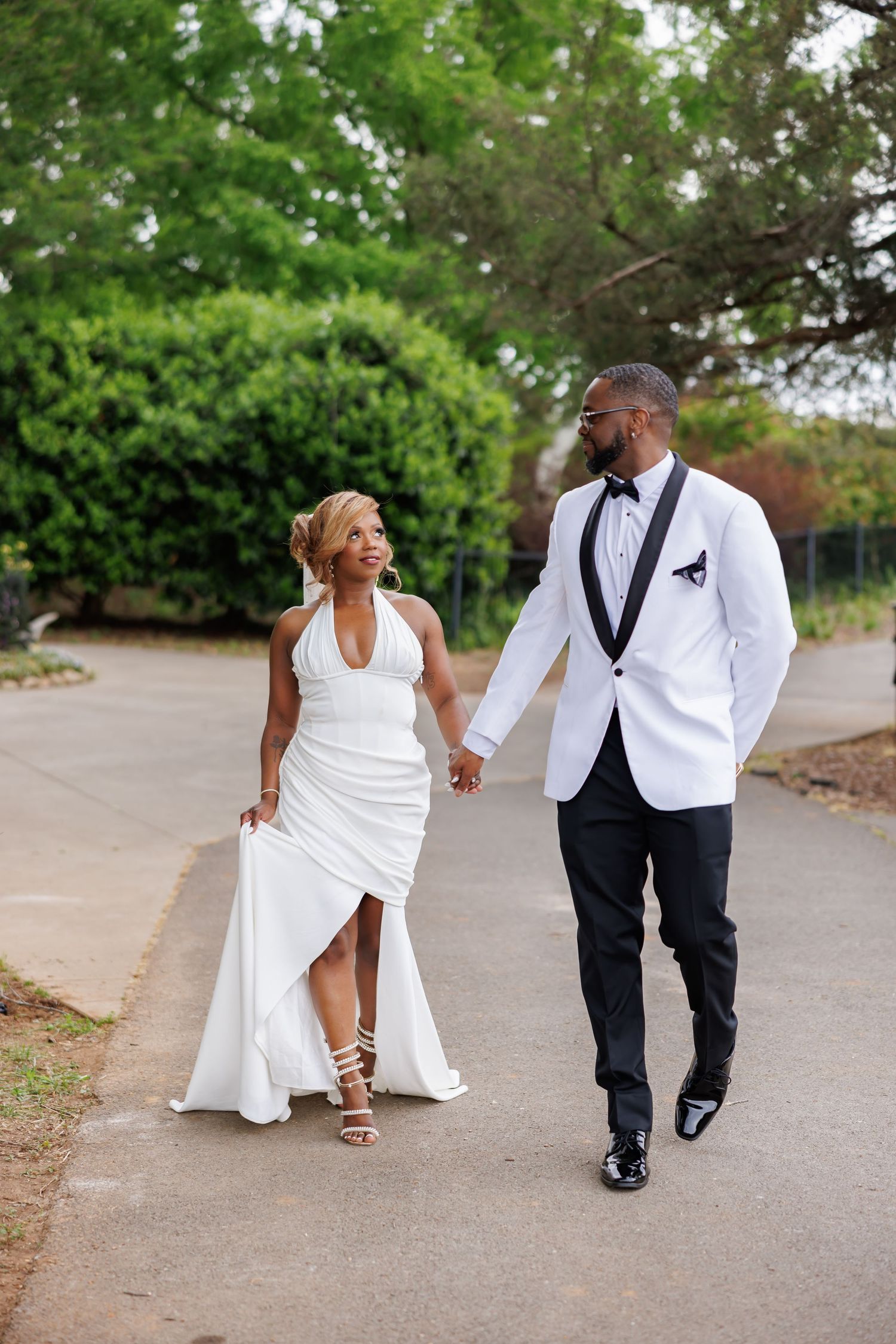 Luxury wedding picture of Bride and groom walk hand in hand in white wedding attire along a tree-lined outdoor pathway.