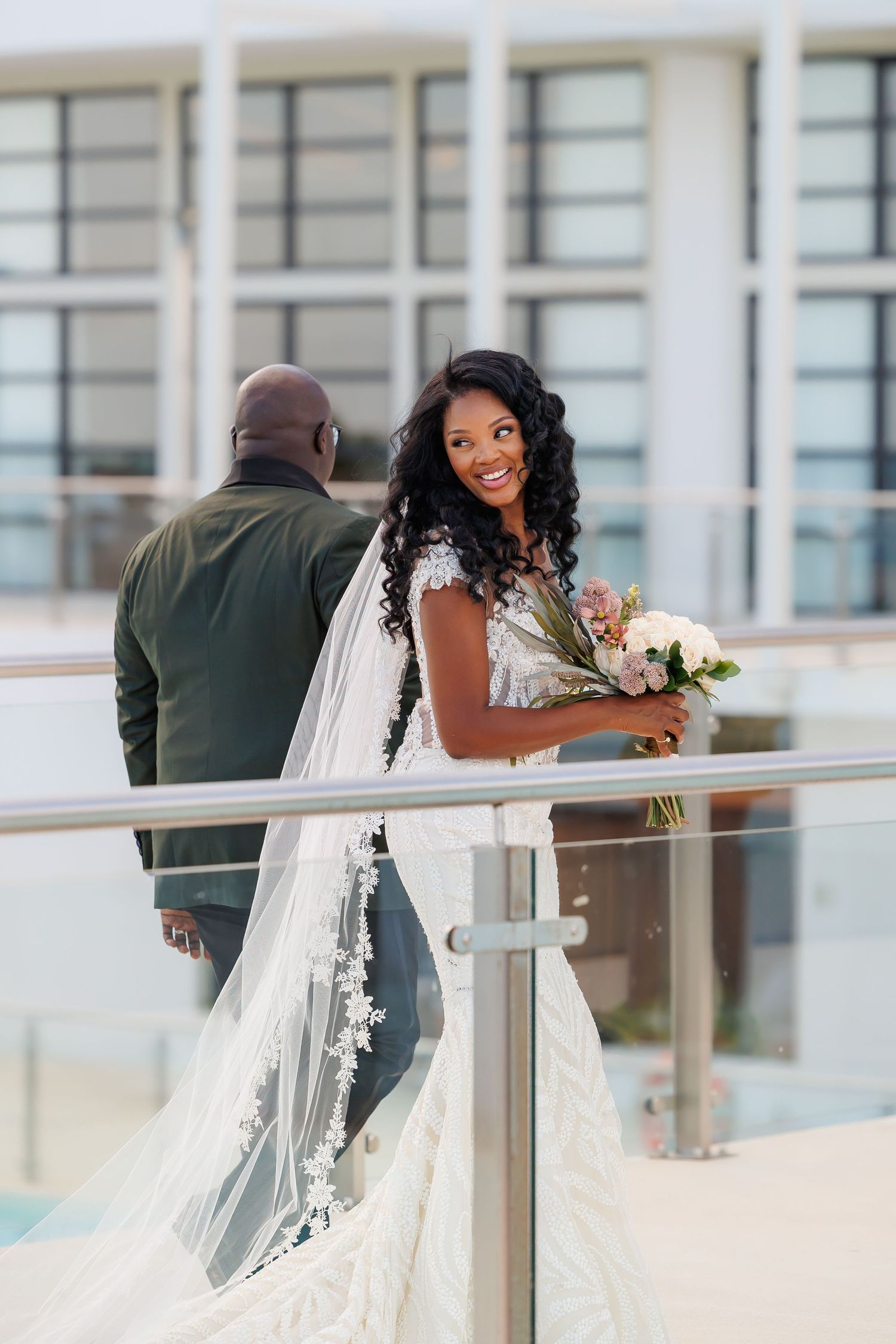 Luxury wedding picture of Radiant bride in lace gown with floral veil holds bouquet while smiling on an outdoor balcony.
