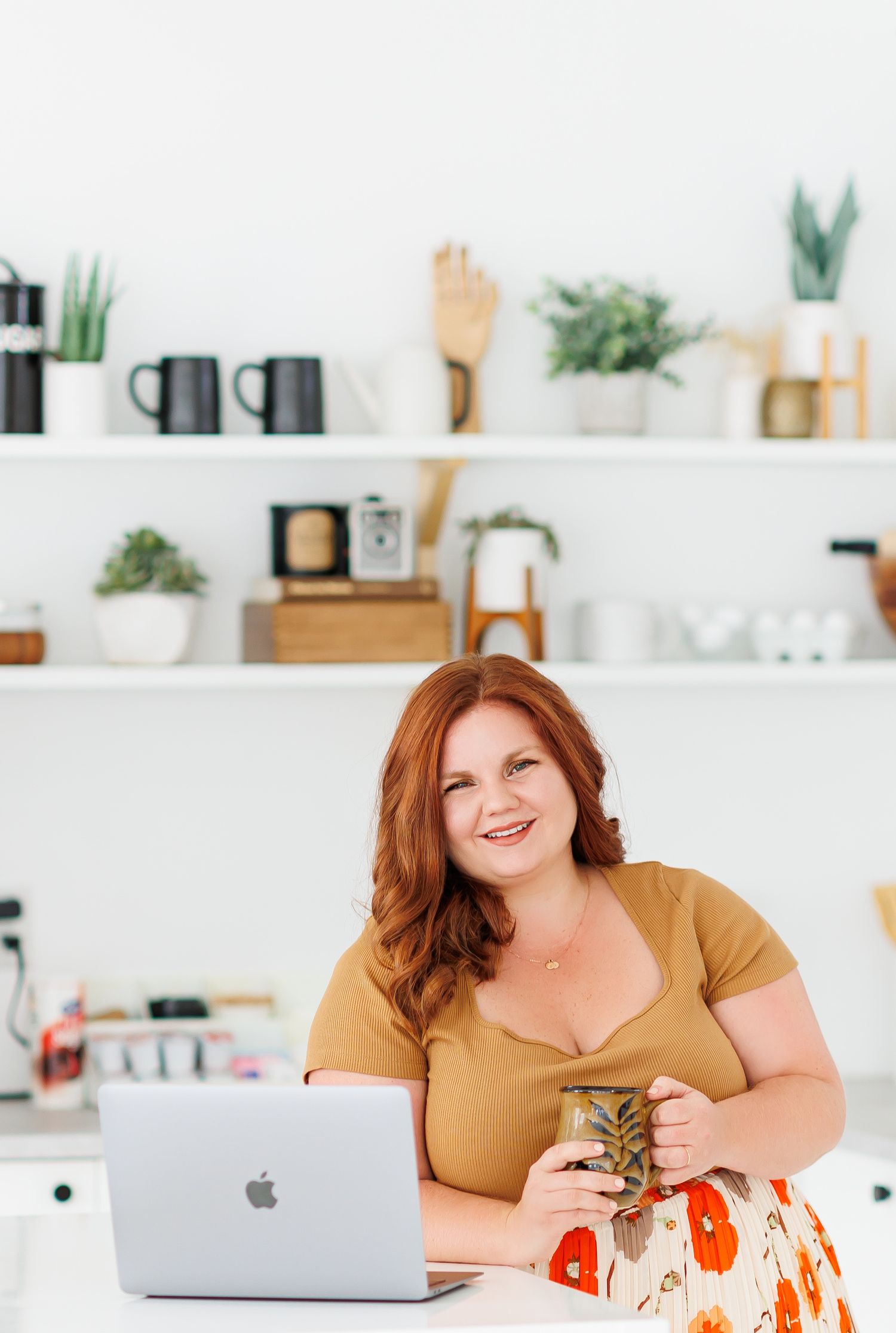 Smiling redhead woman holding a mason jar drink sits at a desk with a MacBook laptop in a bright home office.